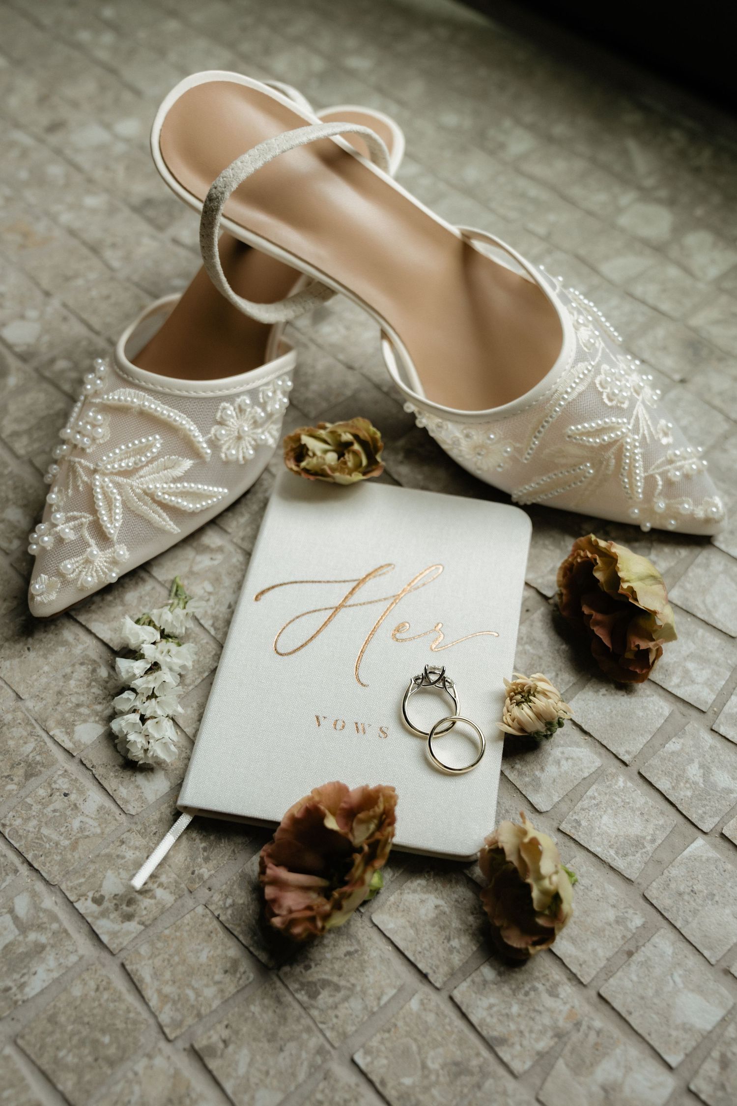 Pearl-embroidered bridal heels beside a Her Vows book, wedding rings, and dried florals on stone floor.