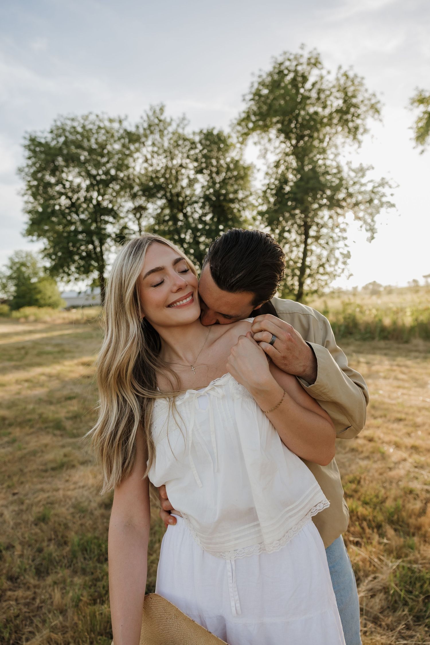 A couple shares a romantic moment outdoors, man kissing woman's shoulder as she smiles, surrounded by green trees.