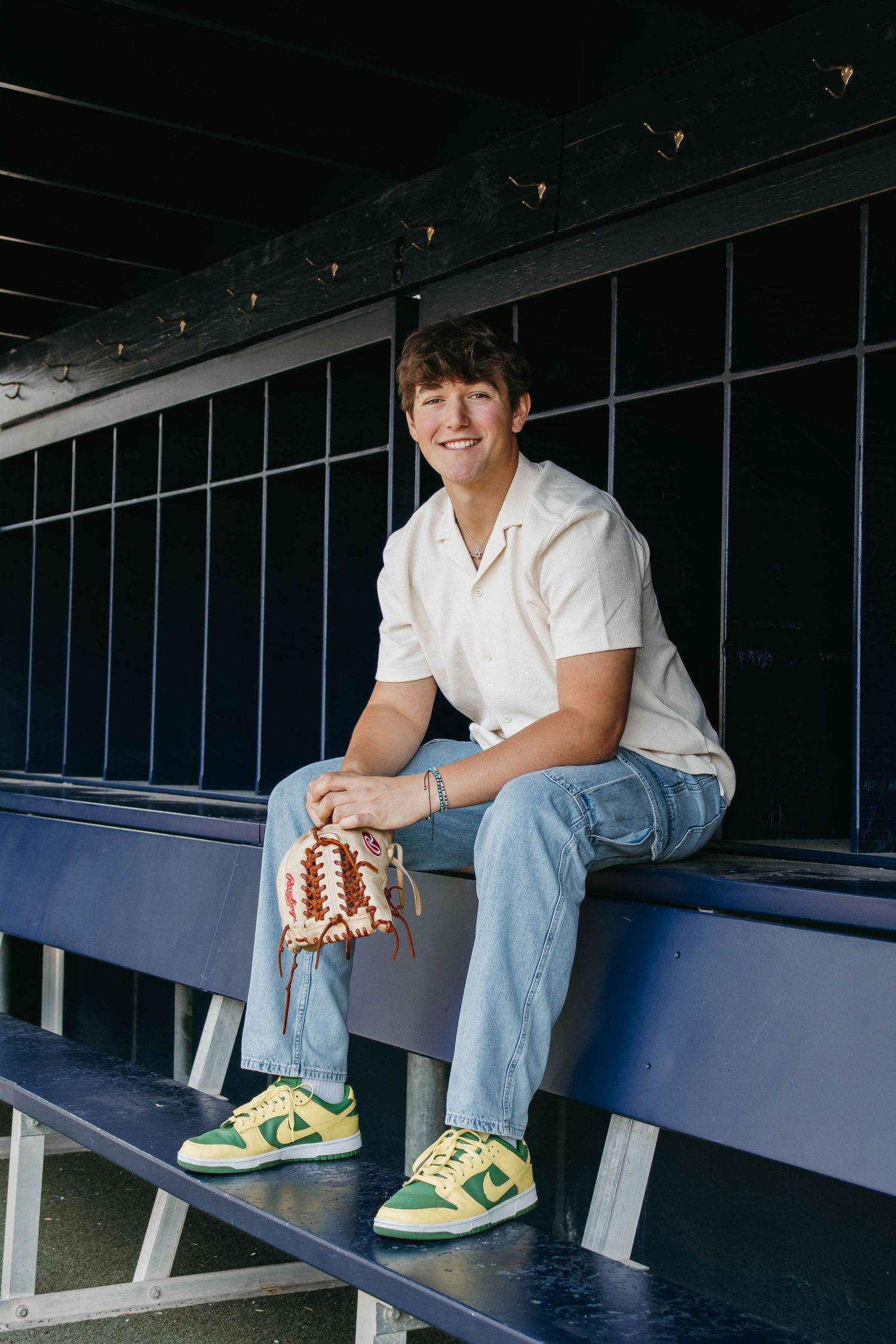 Baseball player in white jersey and jeans sits casually in dugout wearing yellow and green Nike sneakers.
