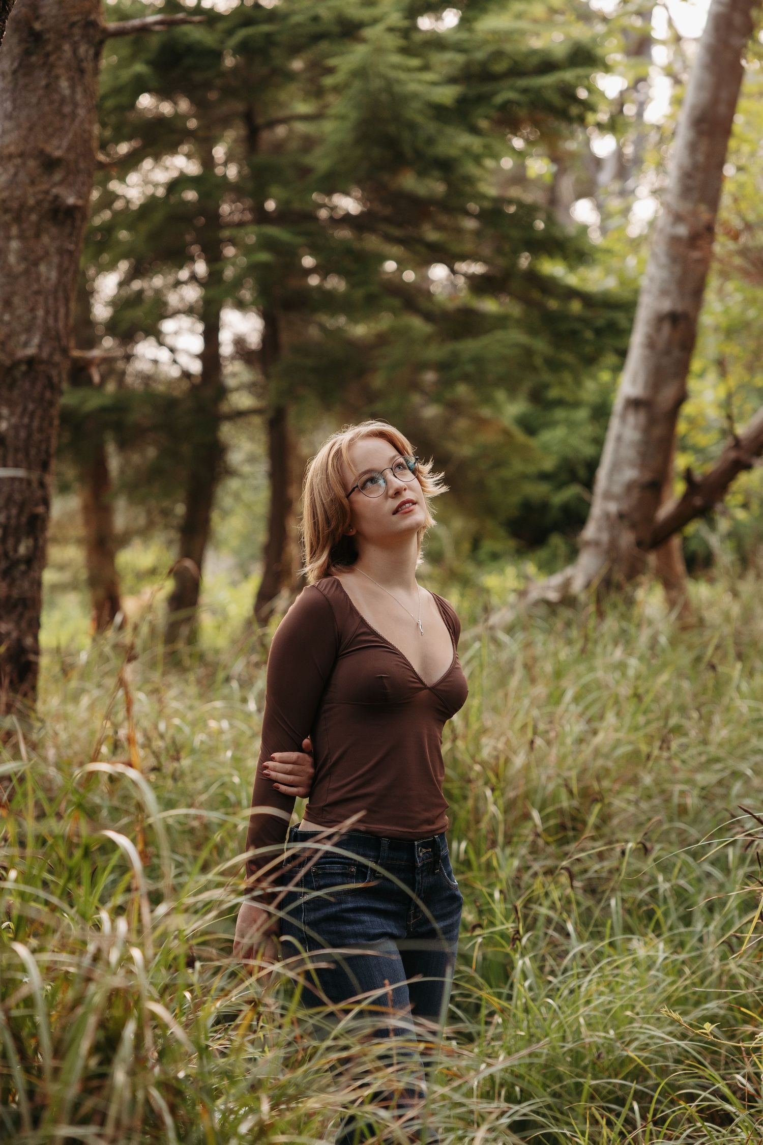 A person in a brown sweater stands thoughtfully among tall grass and trees in a forest setting during golden hour.