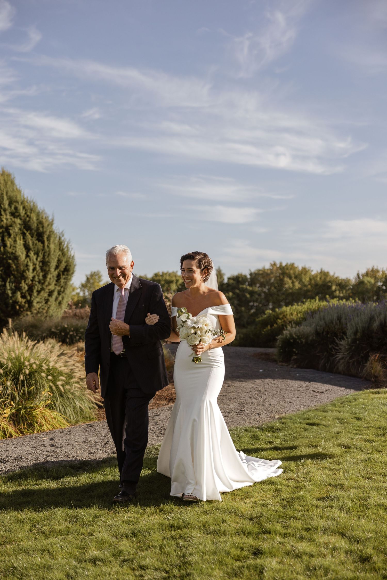 A wedding ceremony processional takes place on a garden path under a bright blue sky.