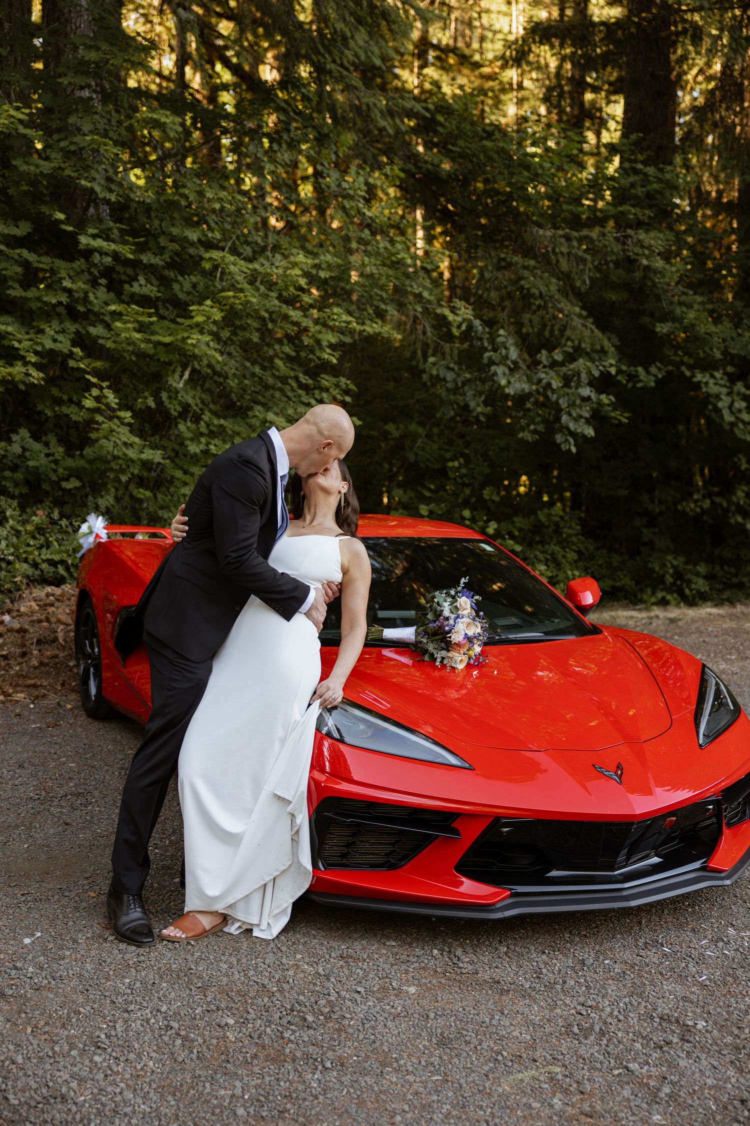 A wedding couple poses romantically against a red Corvette sports car in a forest setting at sunset.