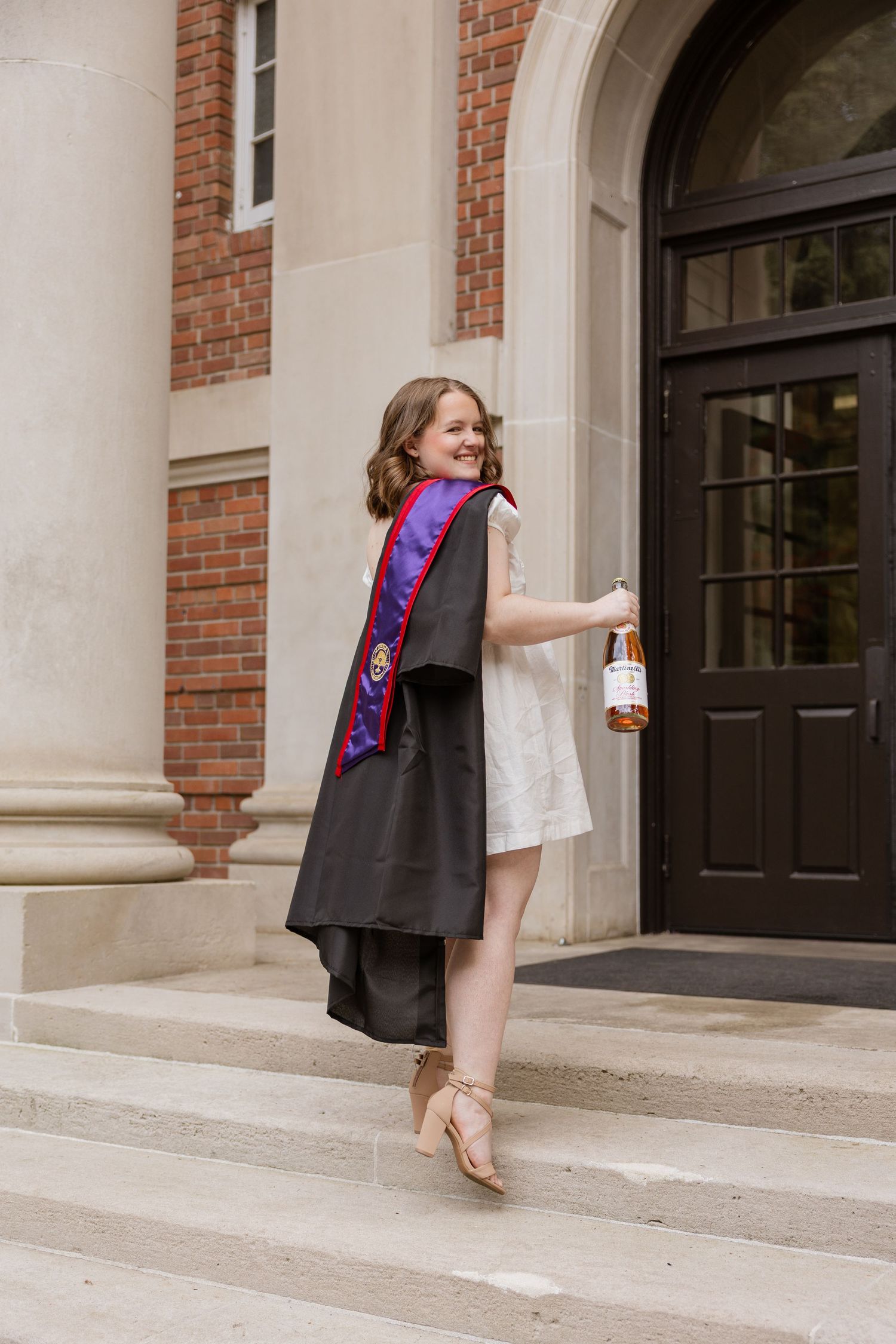 A graduate in academic regalia poses playfully on the steps of a brick building with white stone accents.