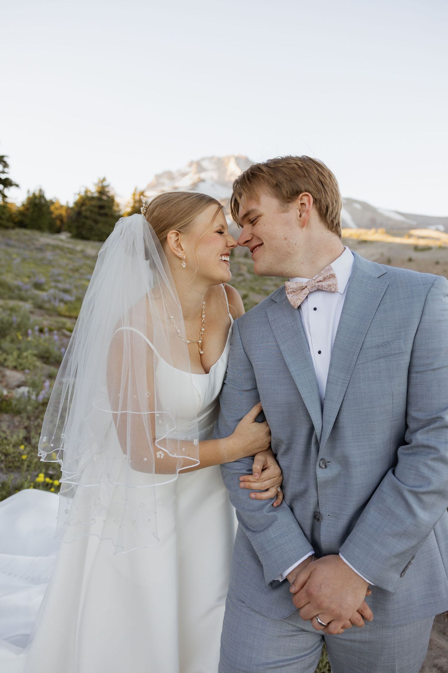 A romantic wedding couple embraces against a mountainous backdrop while wearing elegant grey and white attire.