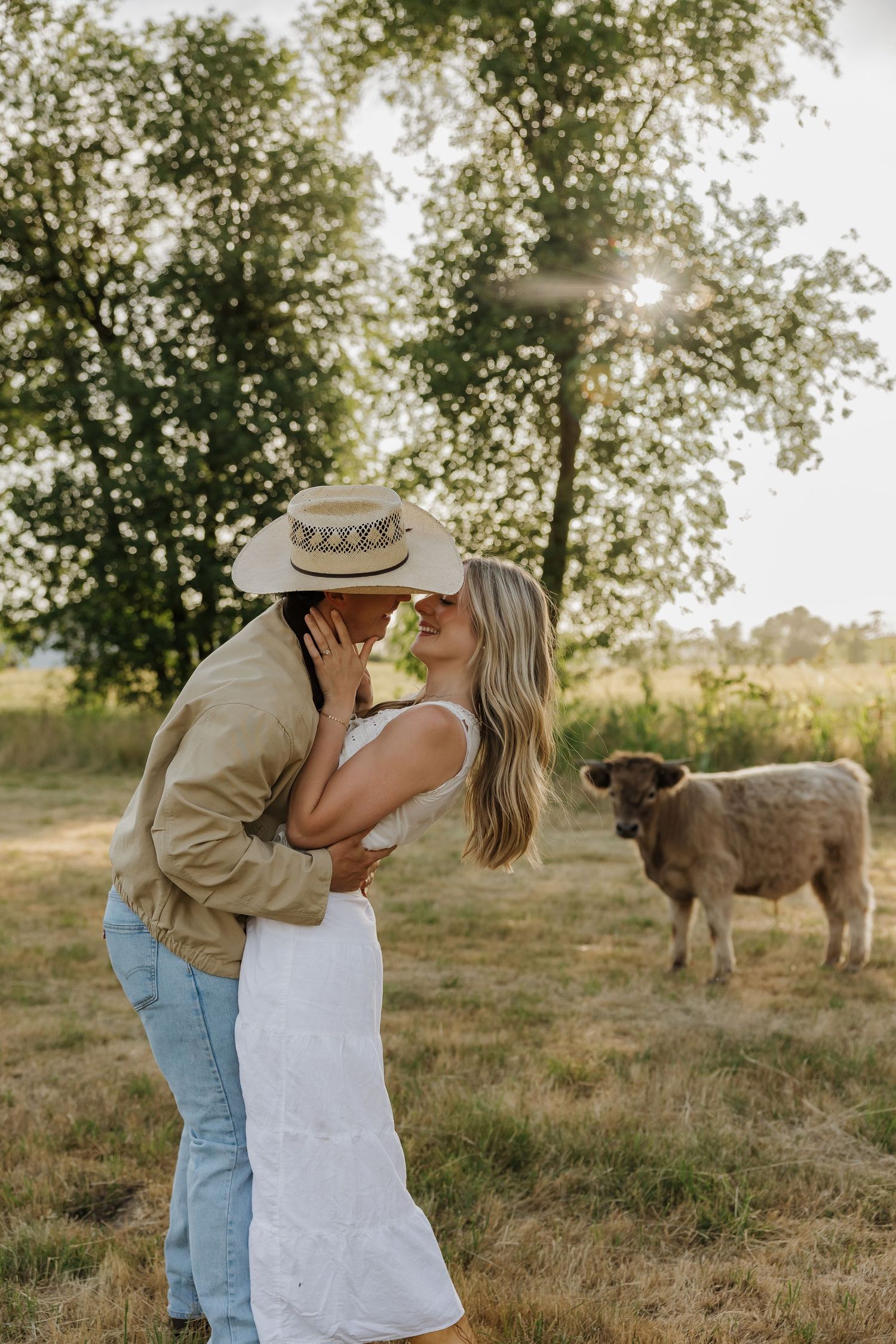 Couple sharing a romantic kiss in a sunlit field with a cow grazing nearby, man wearing cowboy hat and woman in white dress.