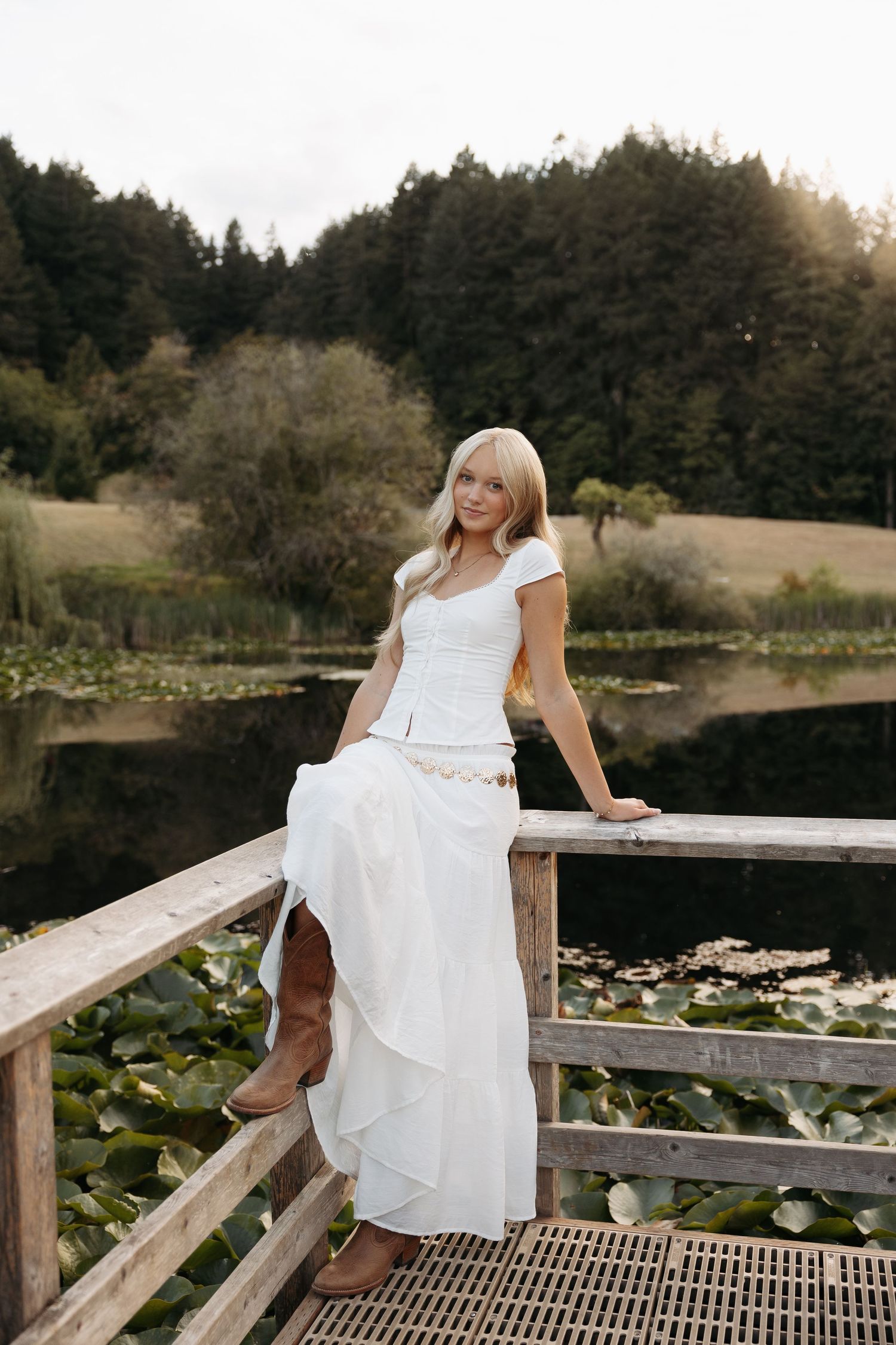 A person in a white wedding dress sits on a wooden railing by a pond with lily pads at sunset.