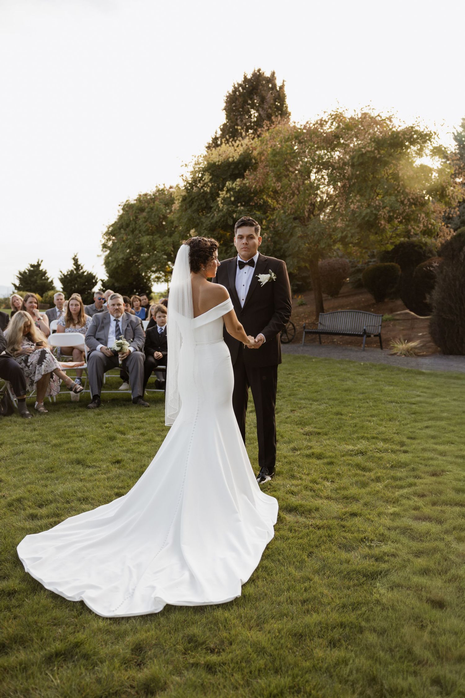 Wedding couple walks across sunlit lawn as guests watch during outdoor evening reception.