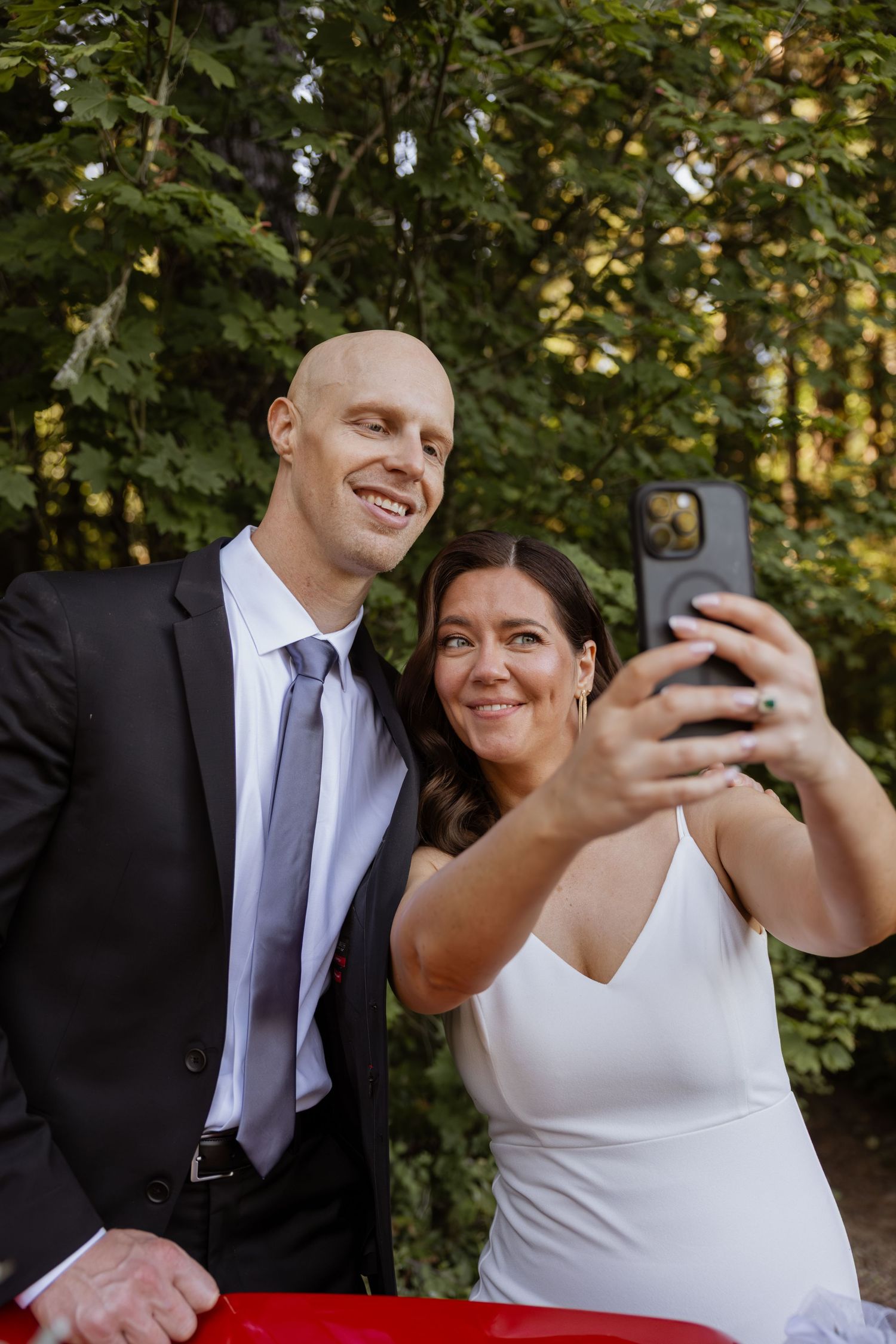 A couple takes selfies together while dressed in formal wedding attire outdoors.