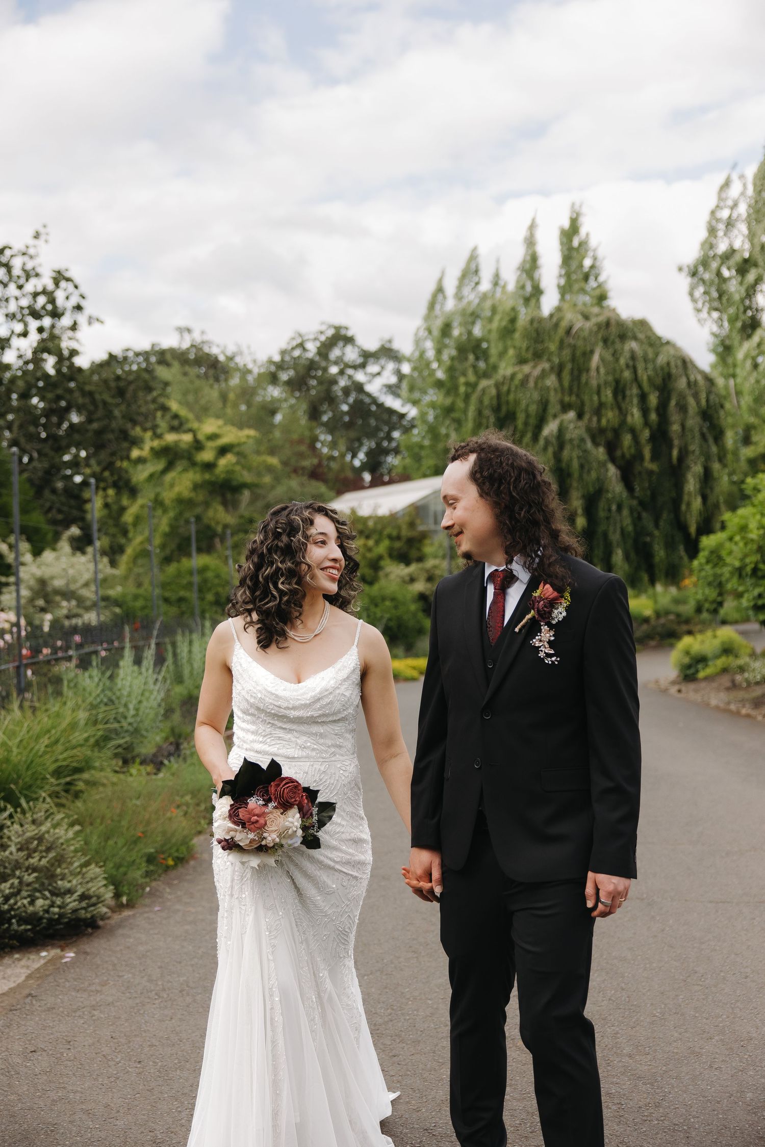 Bride and groom walk hand in hand through a lush garden, smiling at each other on their wedding day.