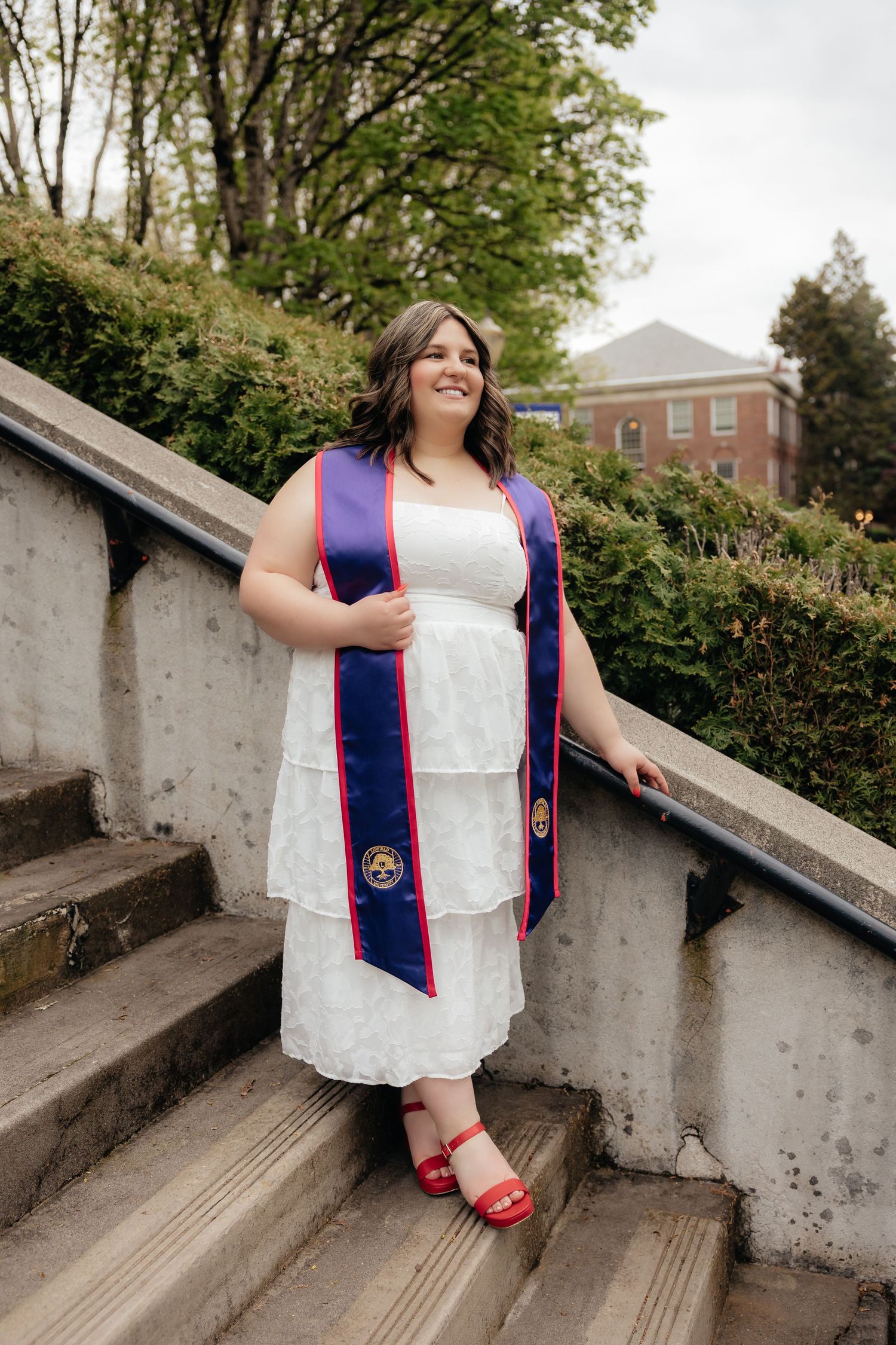 Young woman in white tiered dress and purple graduation stole smiling on stone steps outdoors.