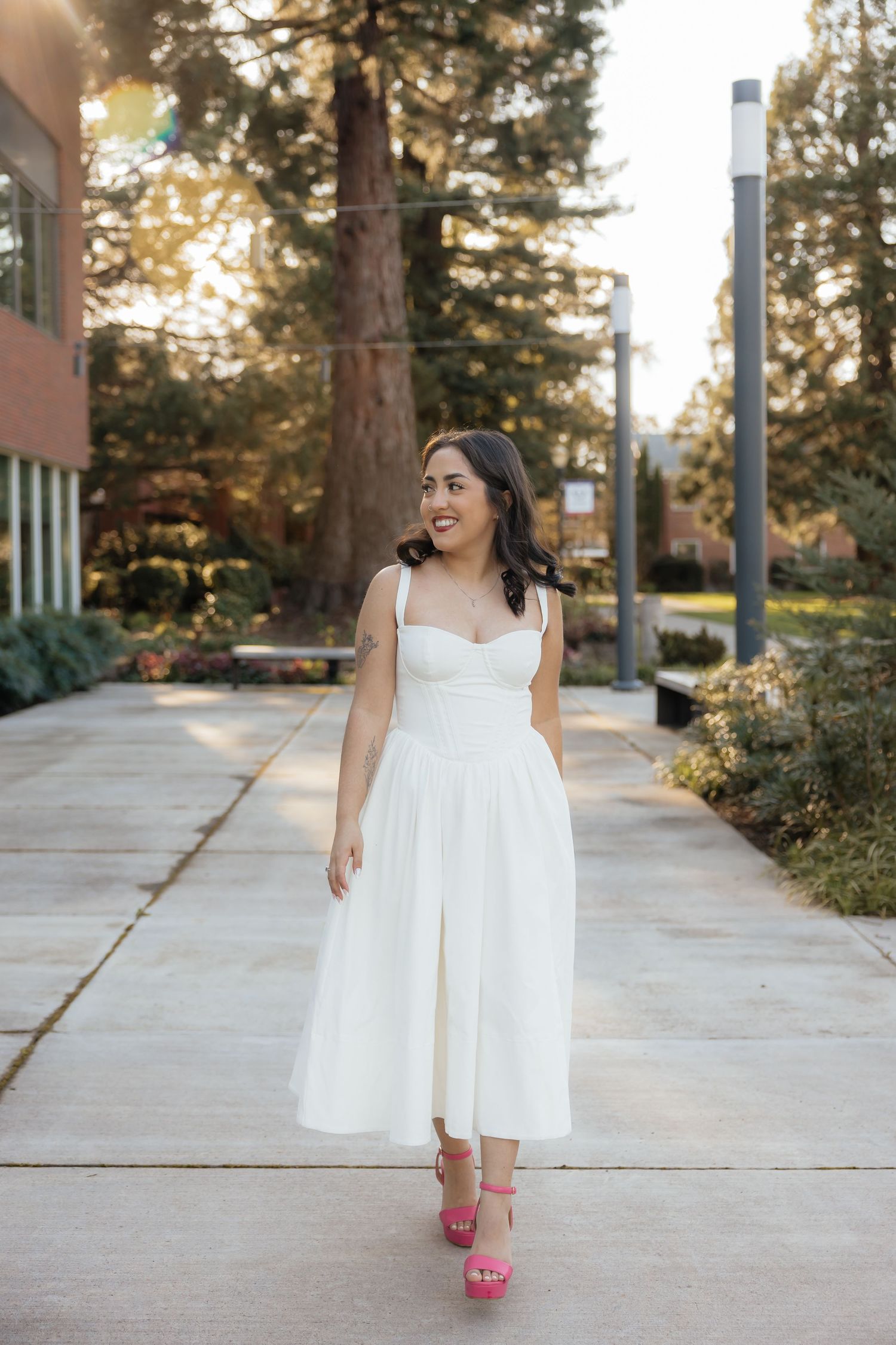 Woman in white midi dress and pink heels walking on a sunny outdoor pathway surrounded by trees.