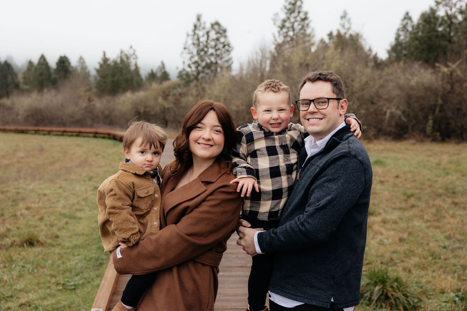 A group of four people pose together outdoors on a wooden boardwalk with evergreen trees in the background.