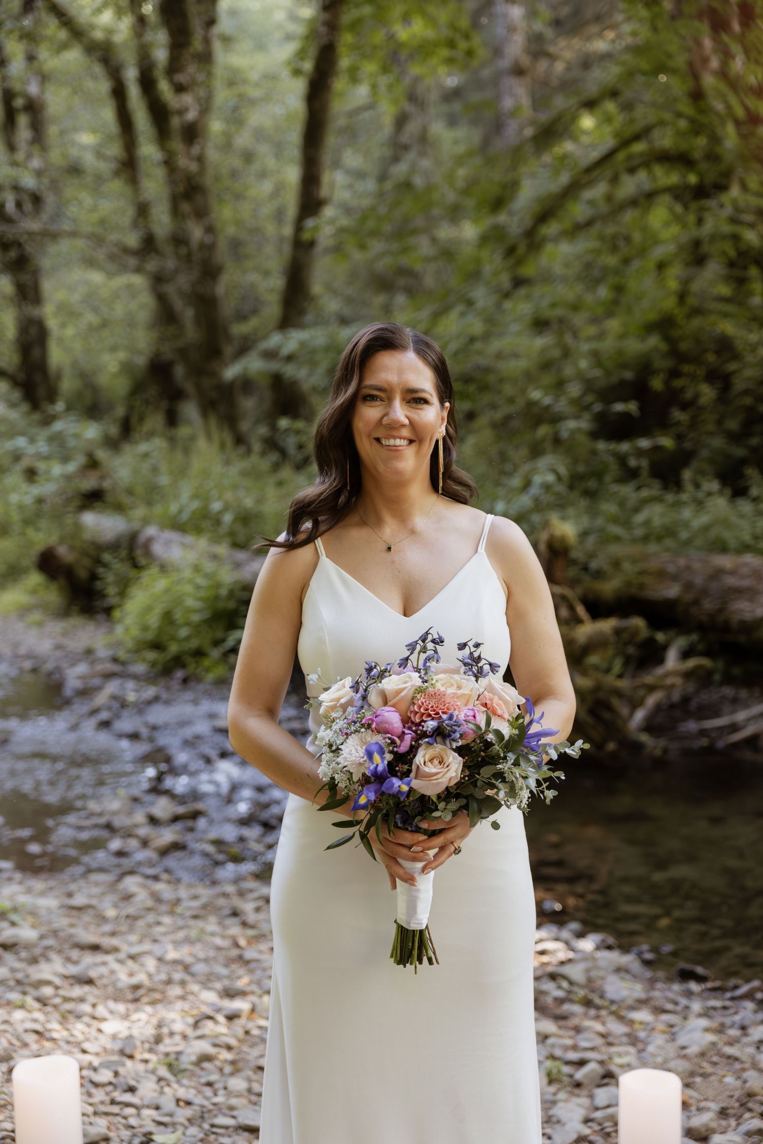 A bride in a white dress holds a purple and pink floral bouquet while standing by a forest stream.
