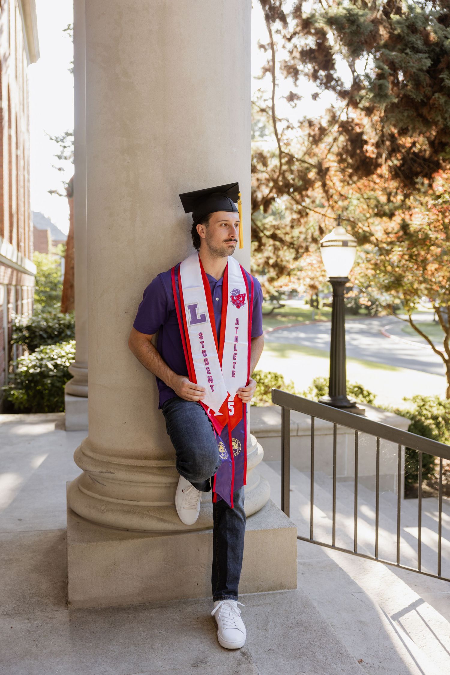 Graduate in purple and white graduation stoles leans against a column while posing for photos on campus.