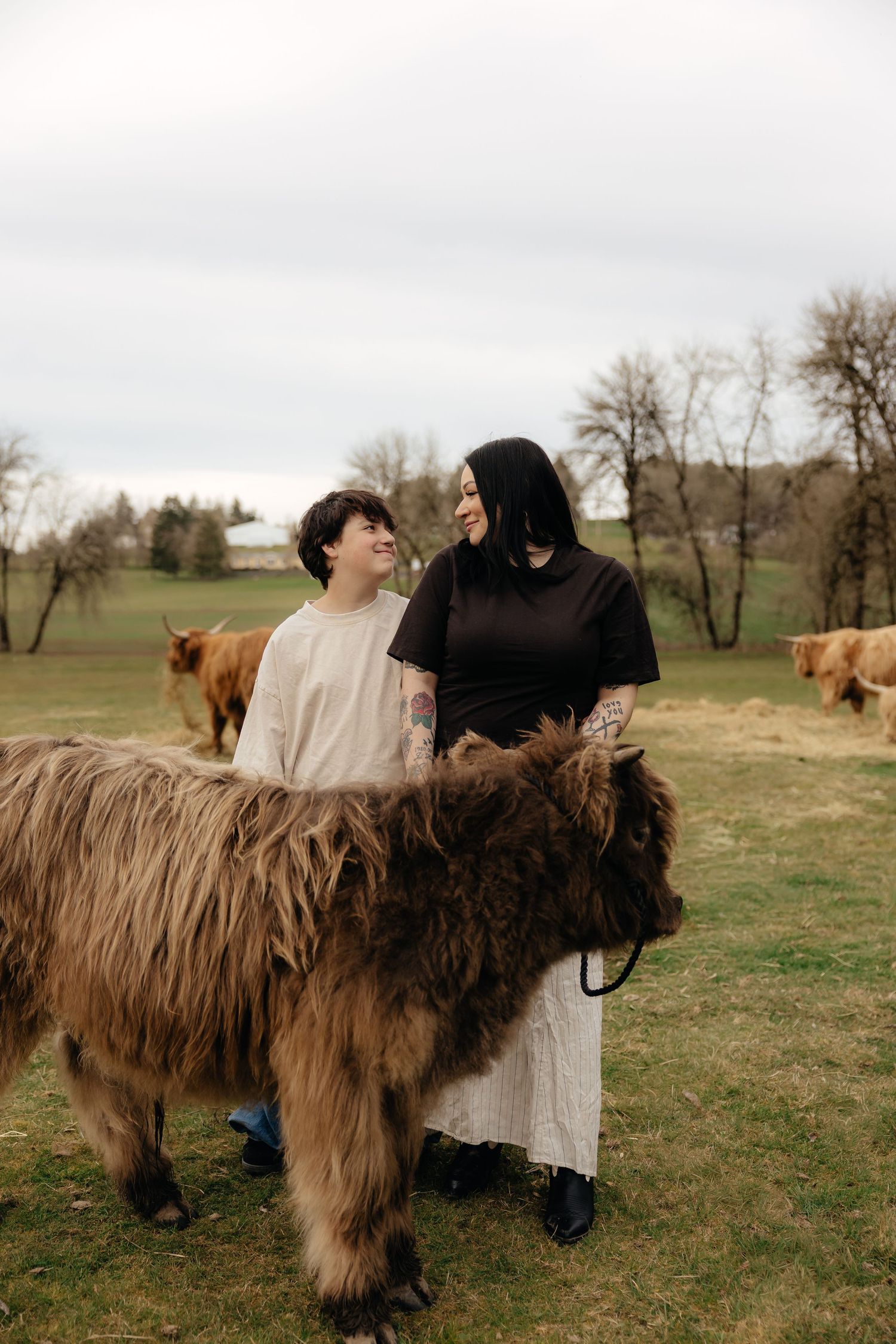 Two people in long dresses stand with a shaggy brown Highland cow in a pastoral field with cattle grazing in background.