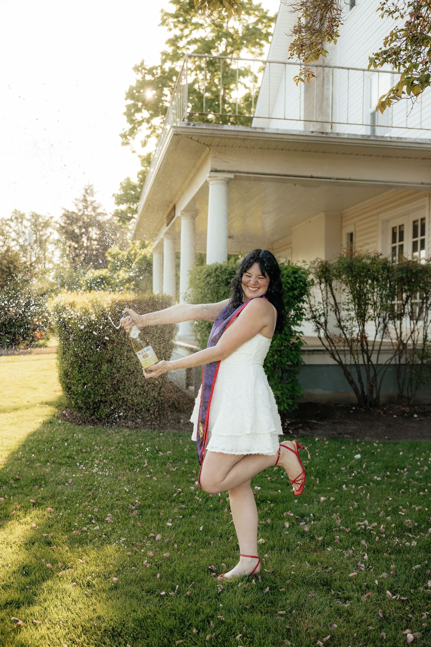 Young woman in white dress popping champagne bottle outdoors on green lawn in front of white colonial house.