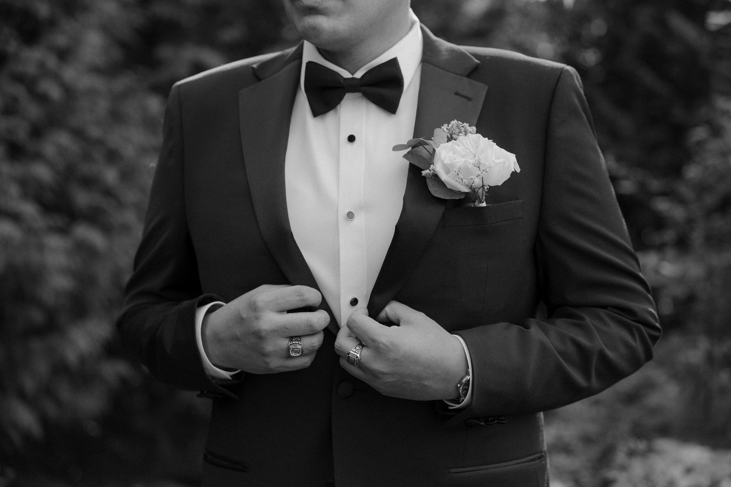 Black and white photo of groomsmen in tuxedos adjusting their bowties and jackets during a formal wedding event.
