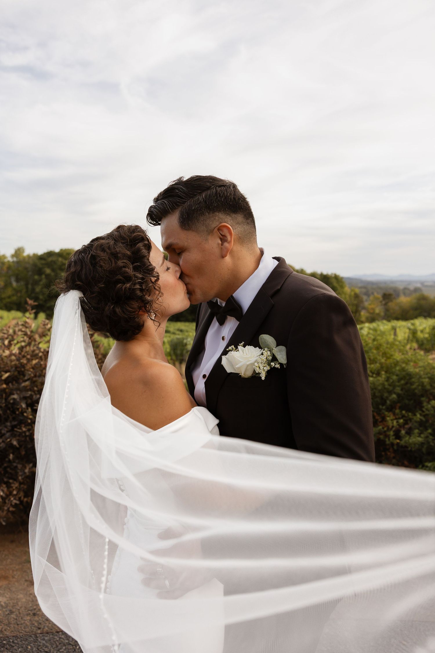 A flowing white wedding veil catches the breeze as newlyweds share an intimate moment outdoors.