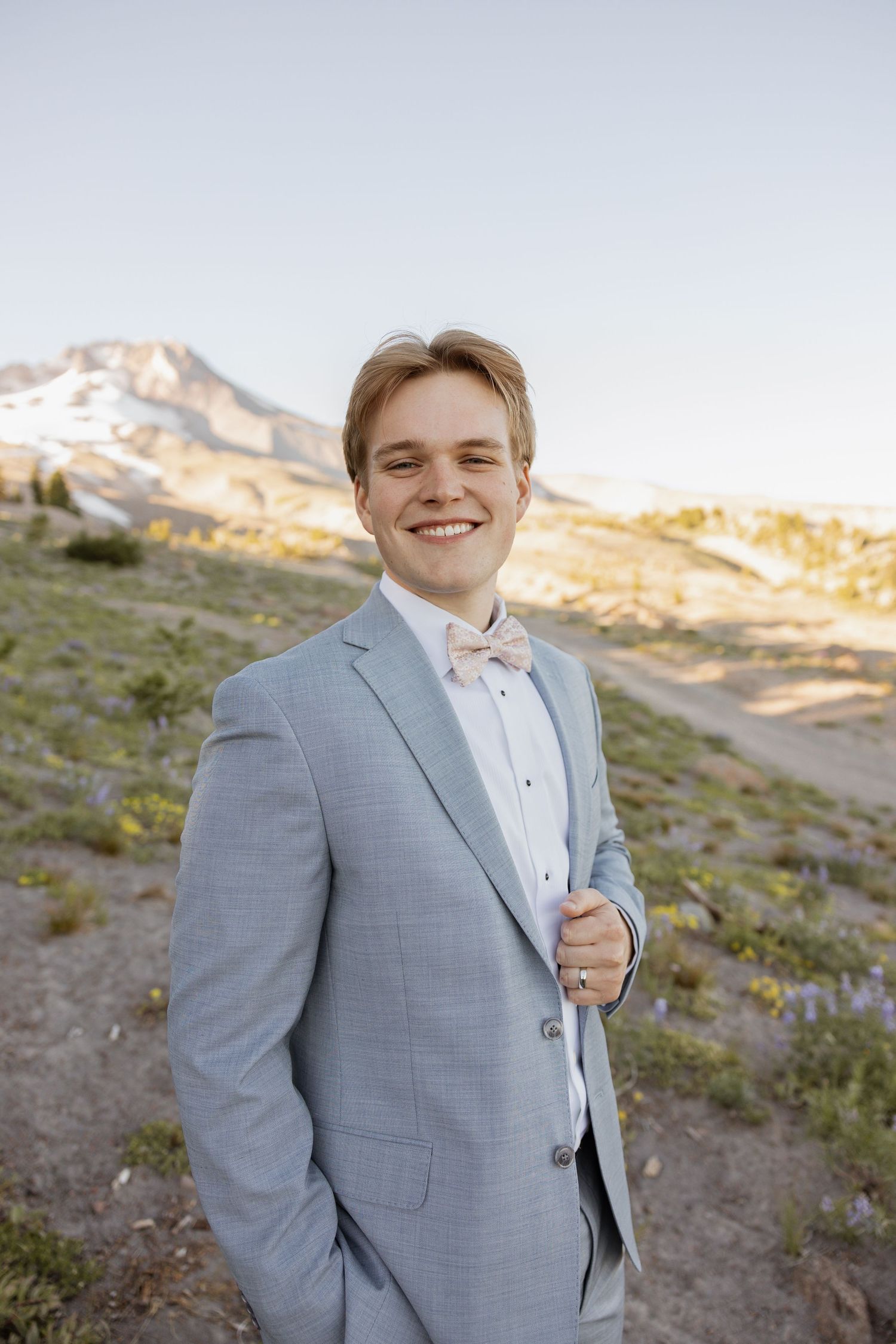 A person in a light blue suit jacket poses outdoors against a rocky mountain landscape at sunset.