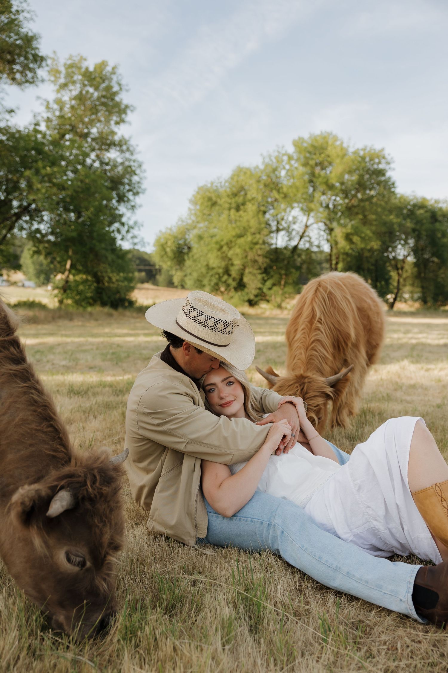 Couple sitting together in a field with a brown calf grazing nearby, man in cowboy hat embracing woman in white dress.