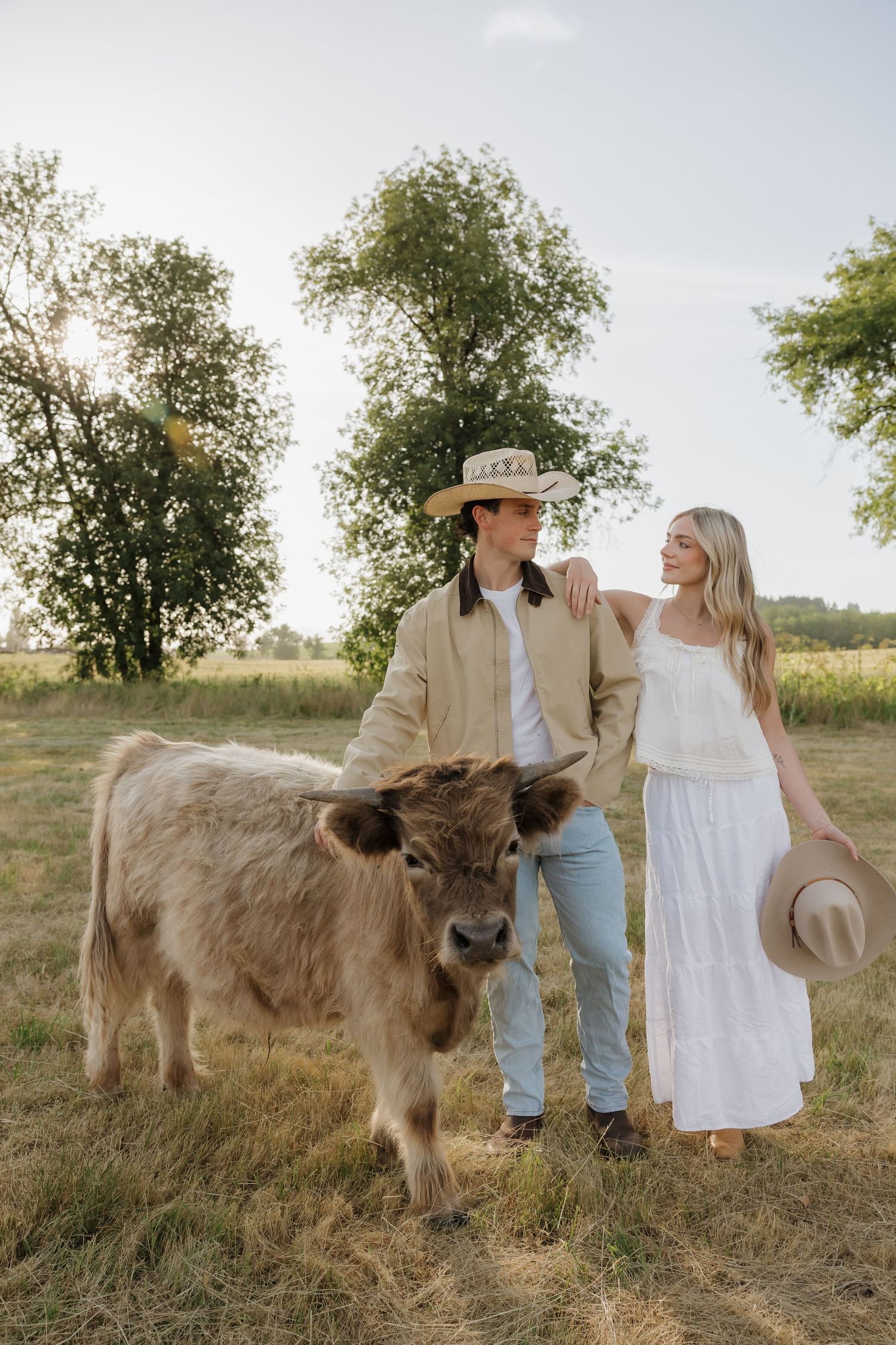 A couple in straw hats stands with a highland calf in a sunny green pasture, the woman in white dress holding her hat.