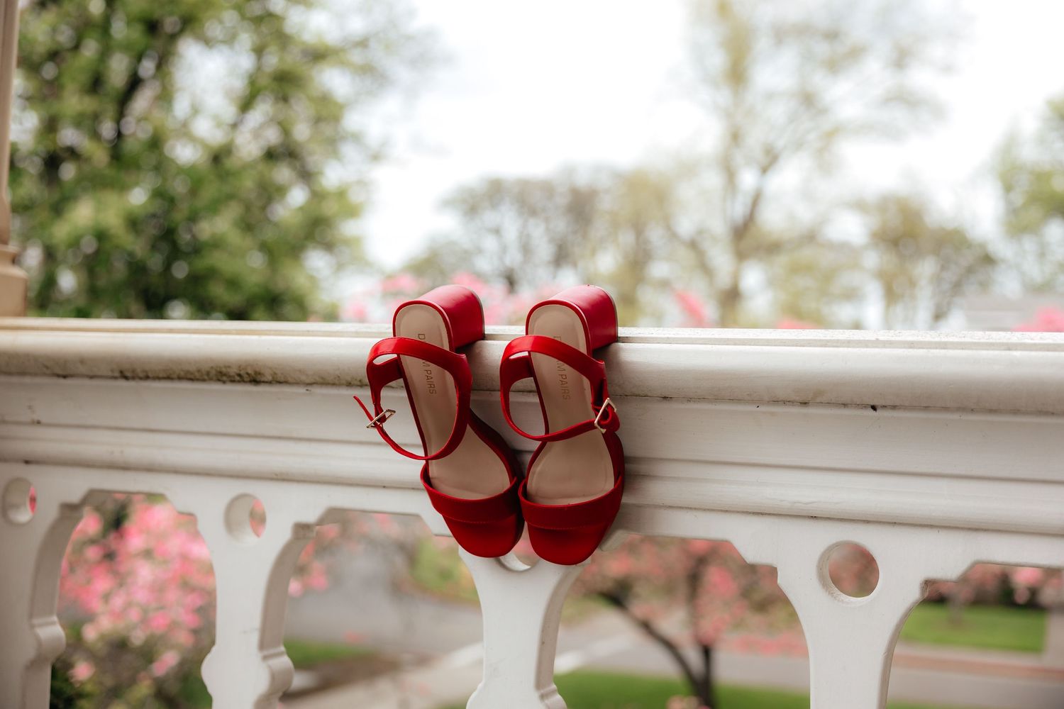 Red strappy heeled sandals balanced on a white balcony railing with blossoming trees in the background.