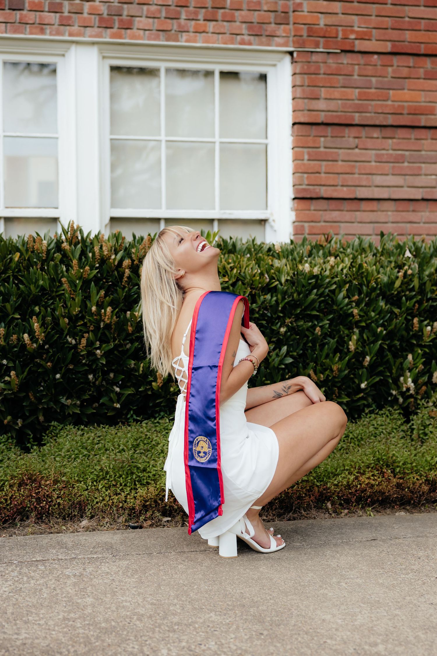 Blonde woman in white dress wearing graduation sash, posing outdoors near brick building and green hedges.