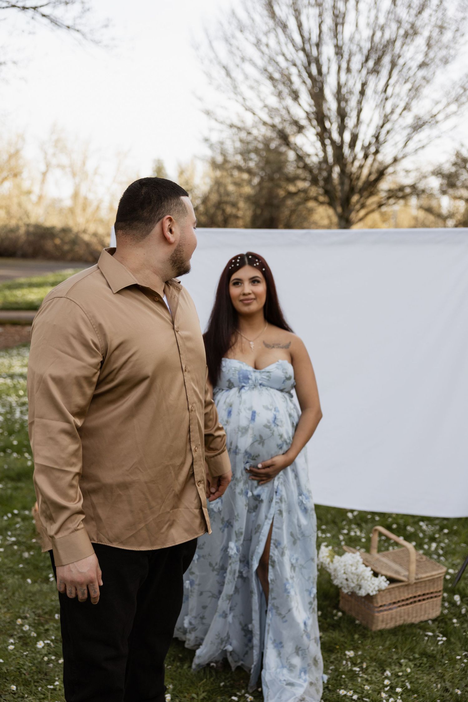 A couple stands together in a spring meadow with white flowers, wearing a tan shirt and blue floral dress.