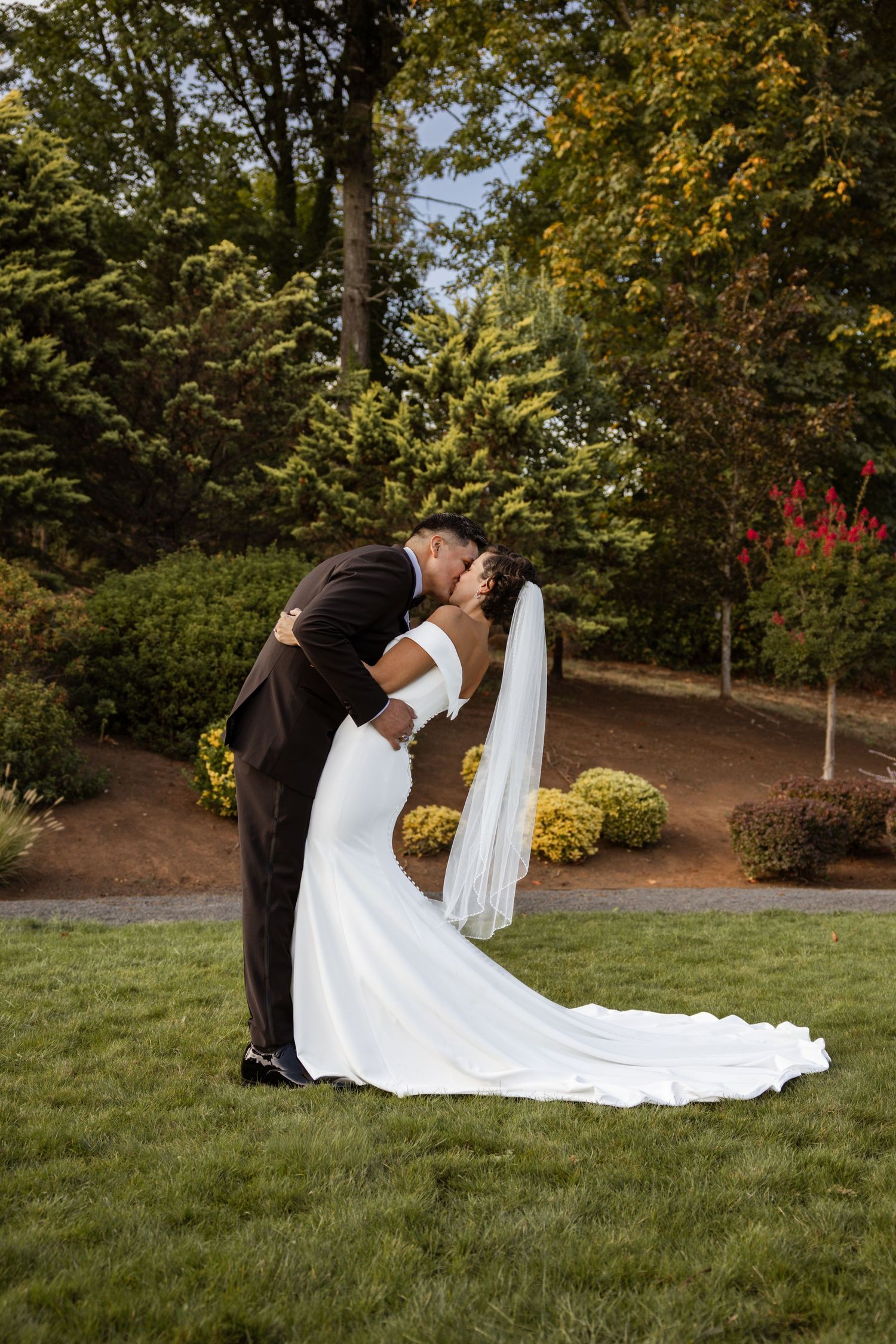 A romantic wedding couple shares a kiss in a lush garden setting while the bride's long white train flows behind them.