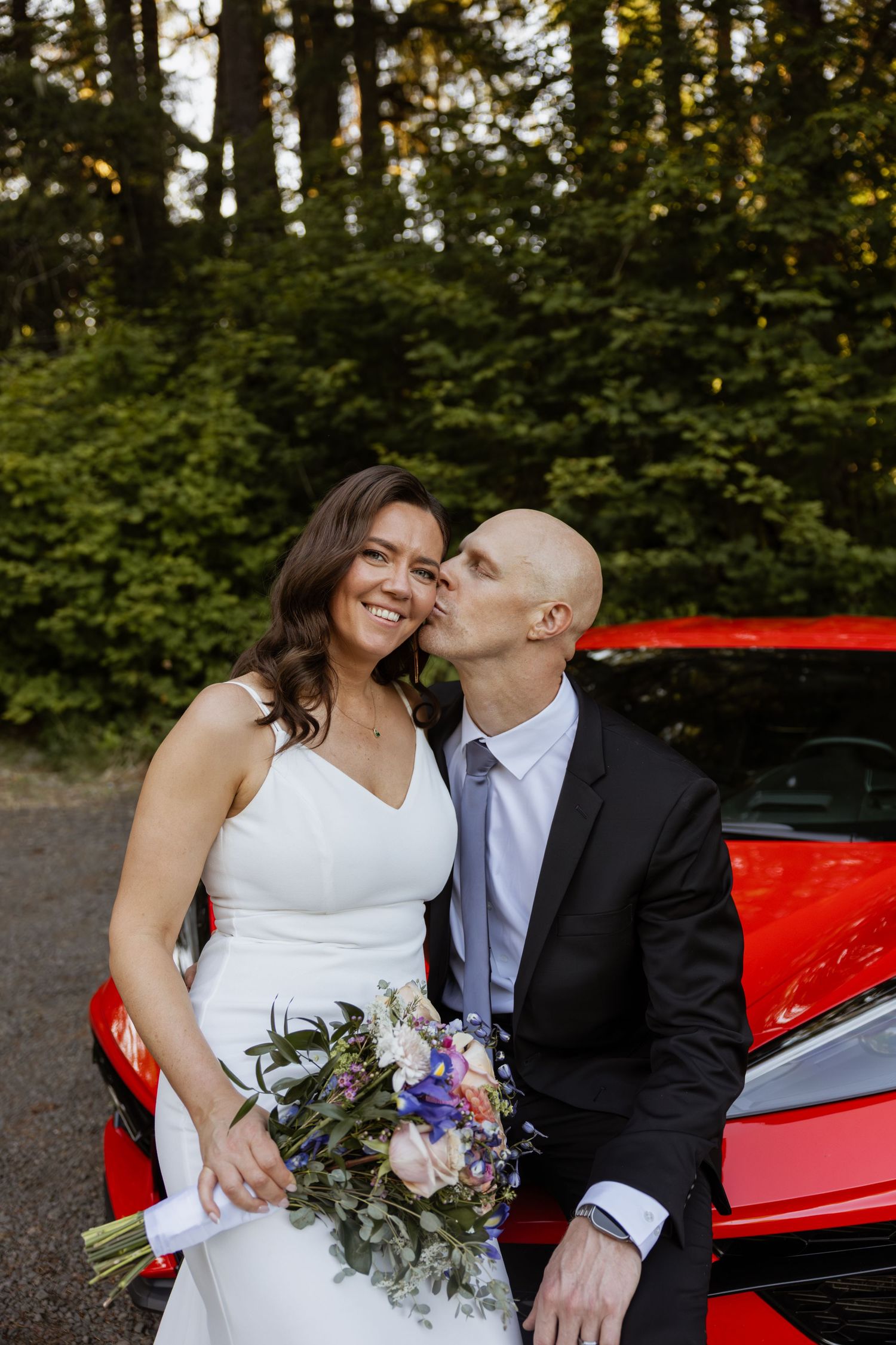 A joyful wedding couple shares a laugh while posing with their red sports car and floral bouquet.