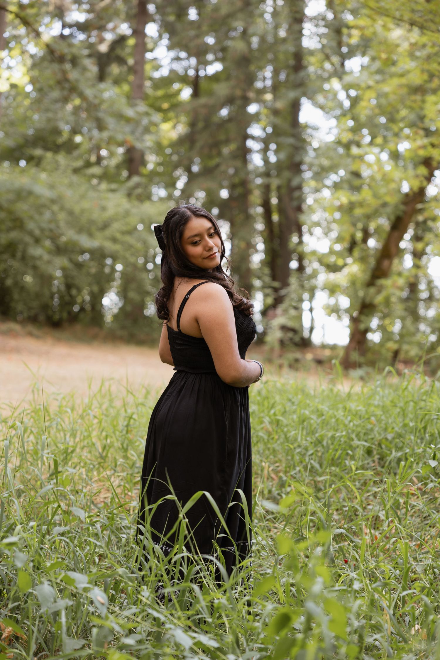 A person in a black dress poses gracefully in a meadow surrounded by tall grass and sunlit trees.