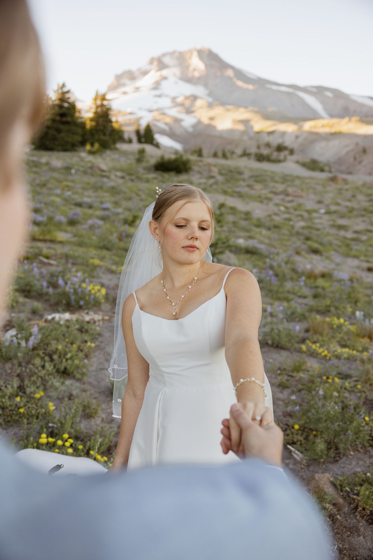A bride in a white wedding dress stands among mountain wildflowers at sunset.