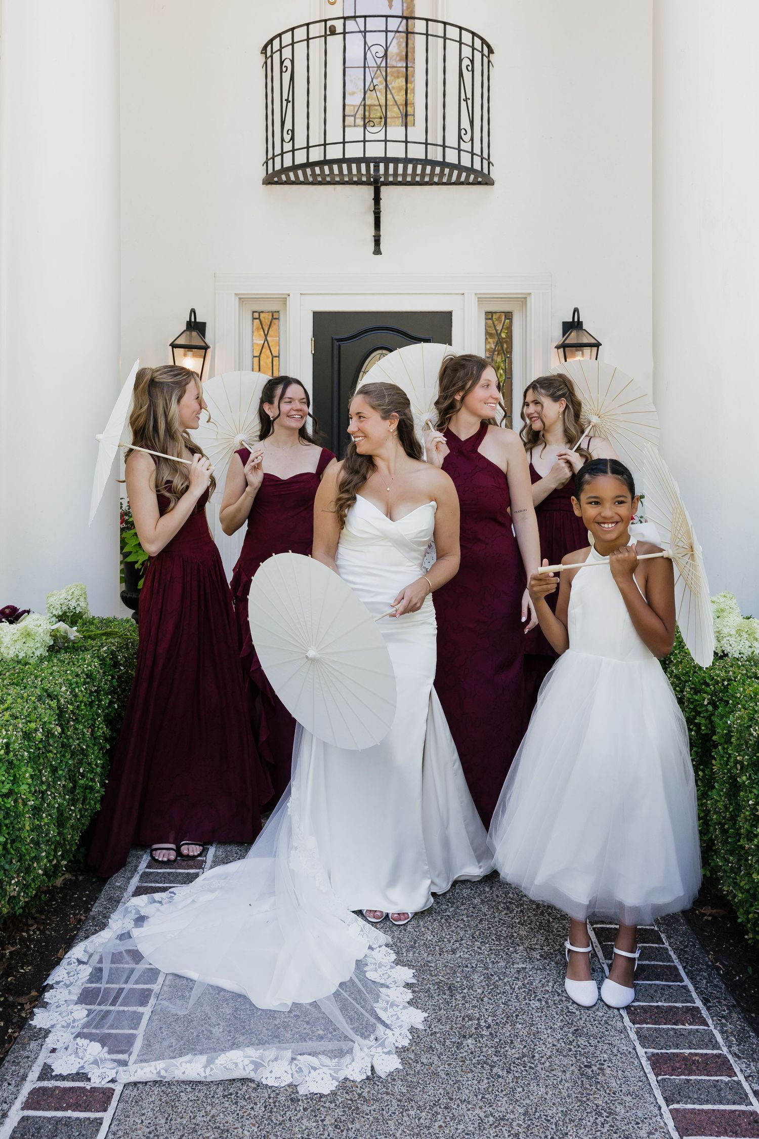 Bride in white gown with bridesmaids in burgundy dresses and flower girl outside elegant venue entrance.