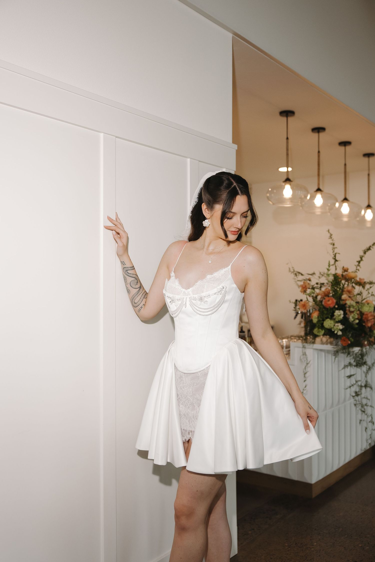 Woman in short white lace bridal mini dress with tattoos poses elegantly in a modern interior setting.