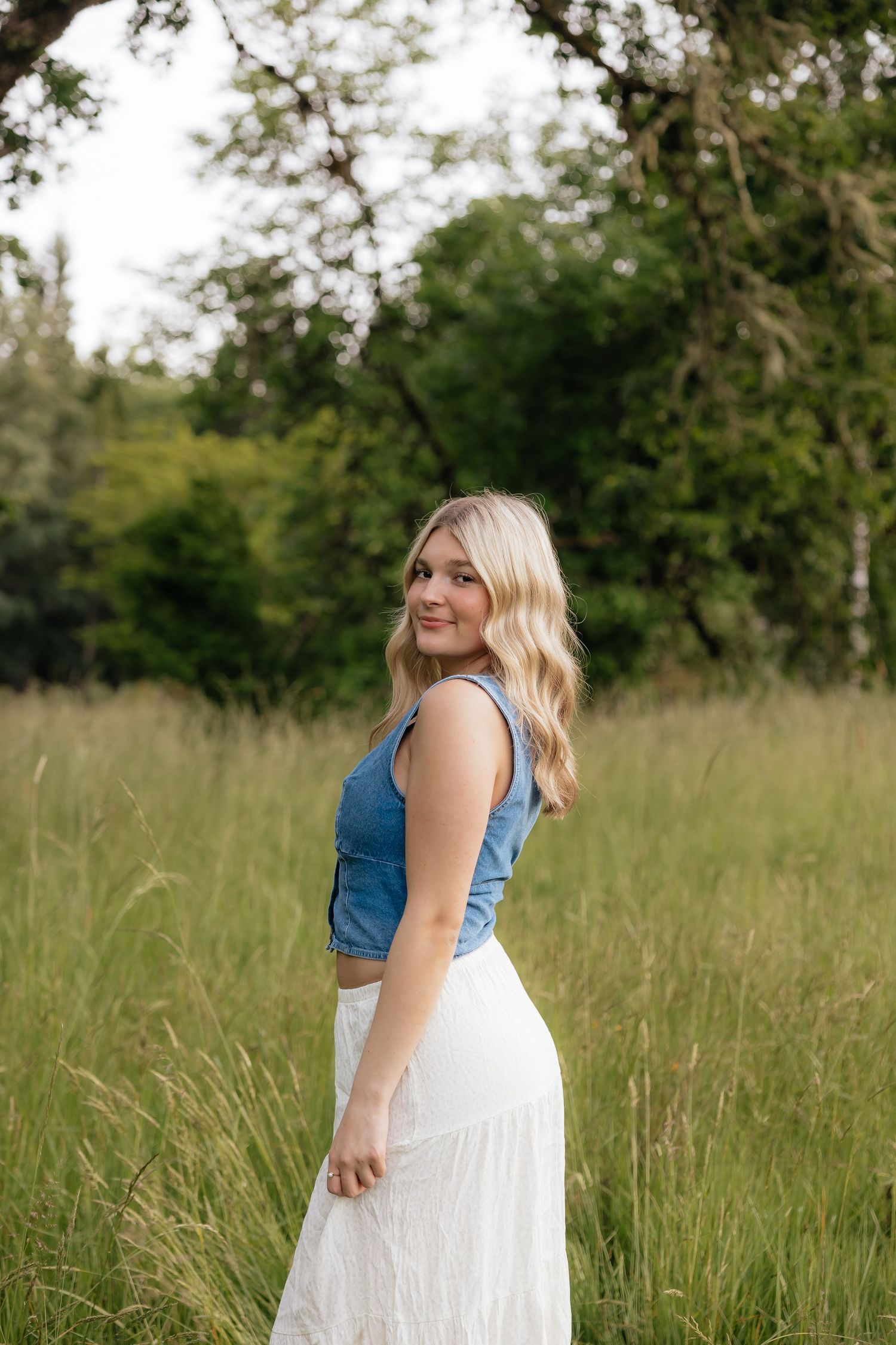 Young blonde woman in denim crop top and white maxi skirt smiling in a lush green meadow.
