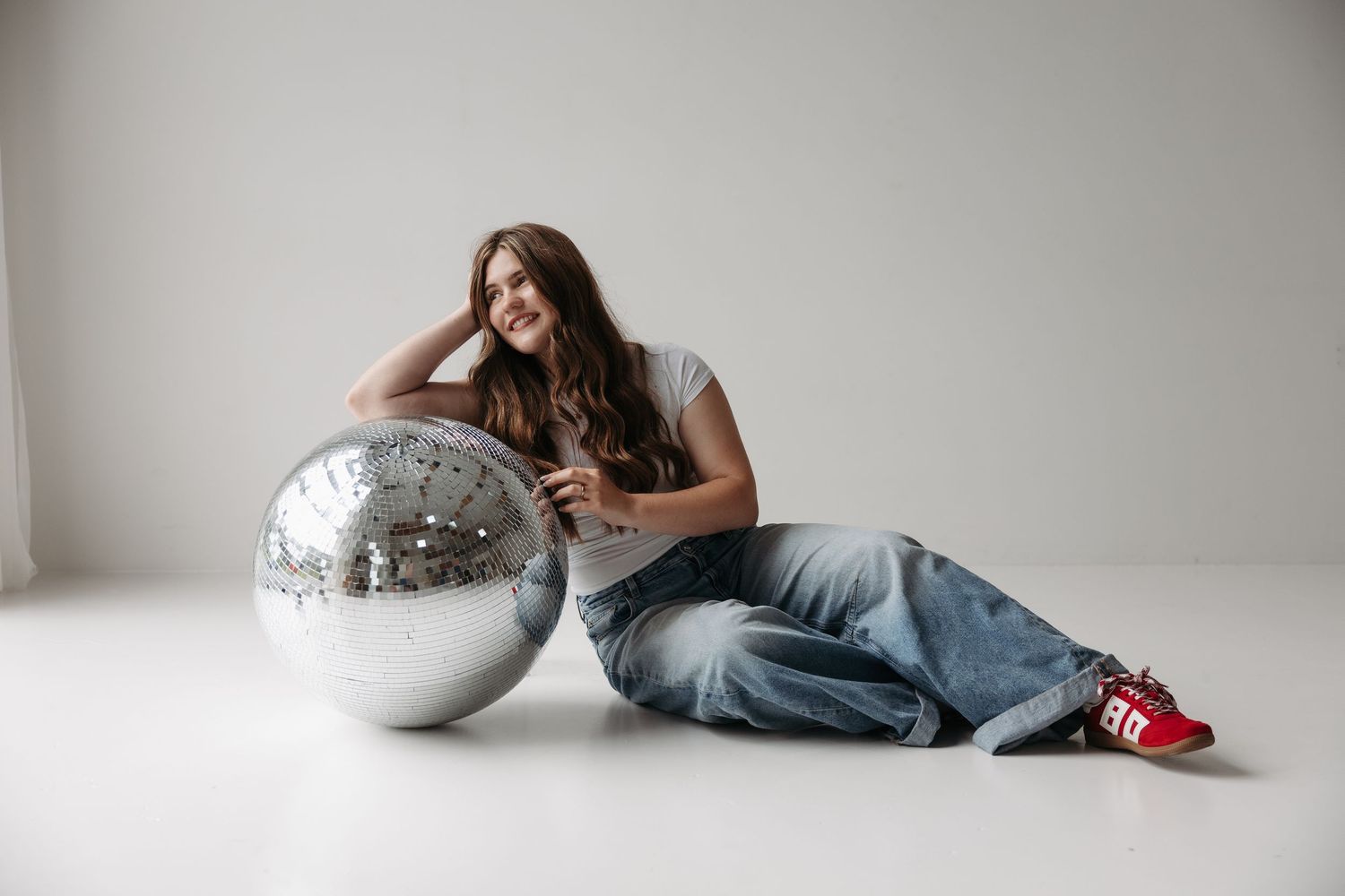 Young woman in jeans and red sneakers sitting on floor leaning against large silver disco ball, smiling.