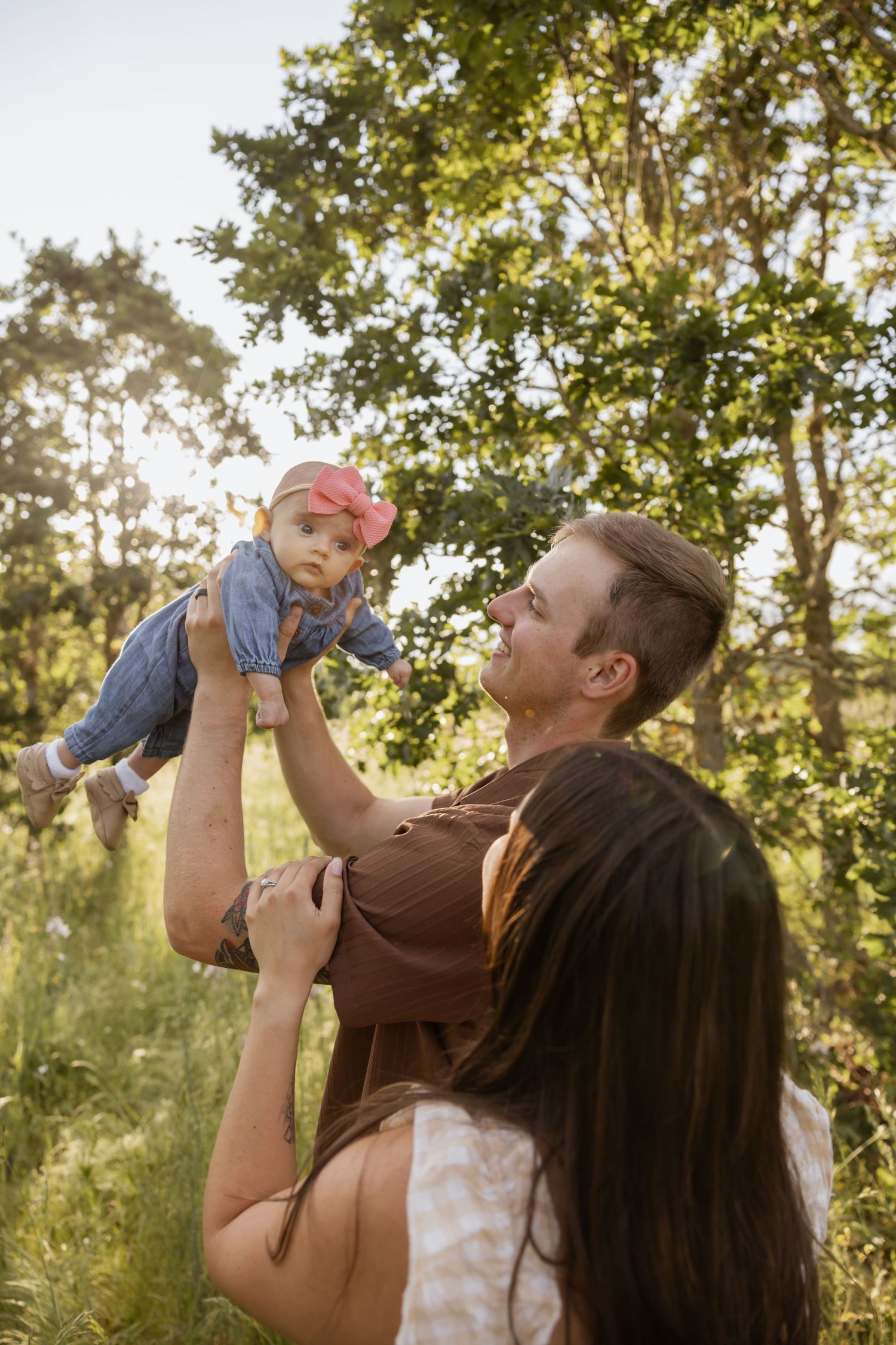 Parents playing with baby outdoors in sunlit forest during golden hour family photoshoot.