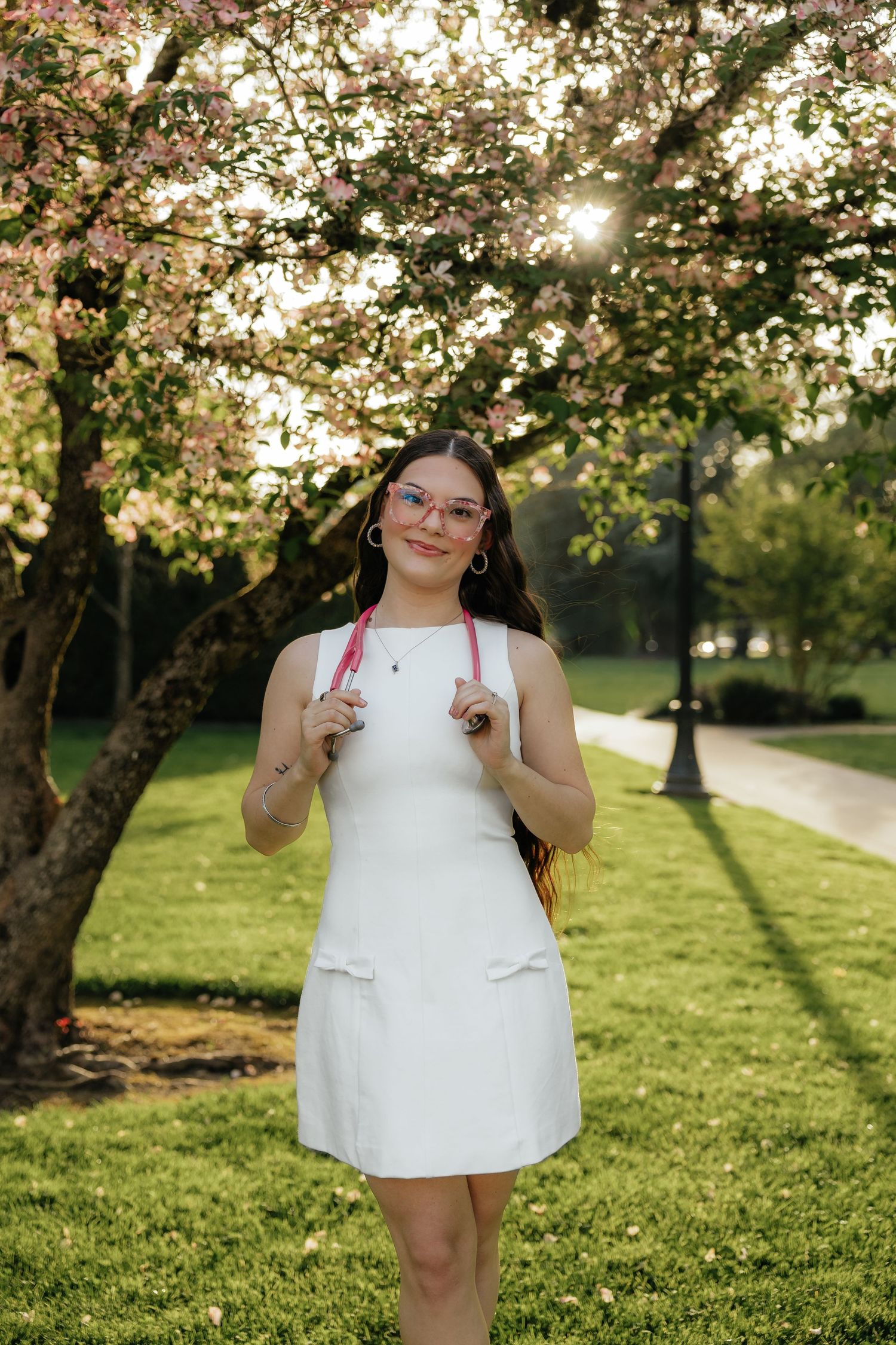Young woman in white dress poses under blooming pink cherry blossom tree in sunny green park.