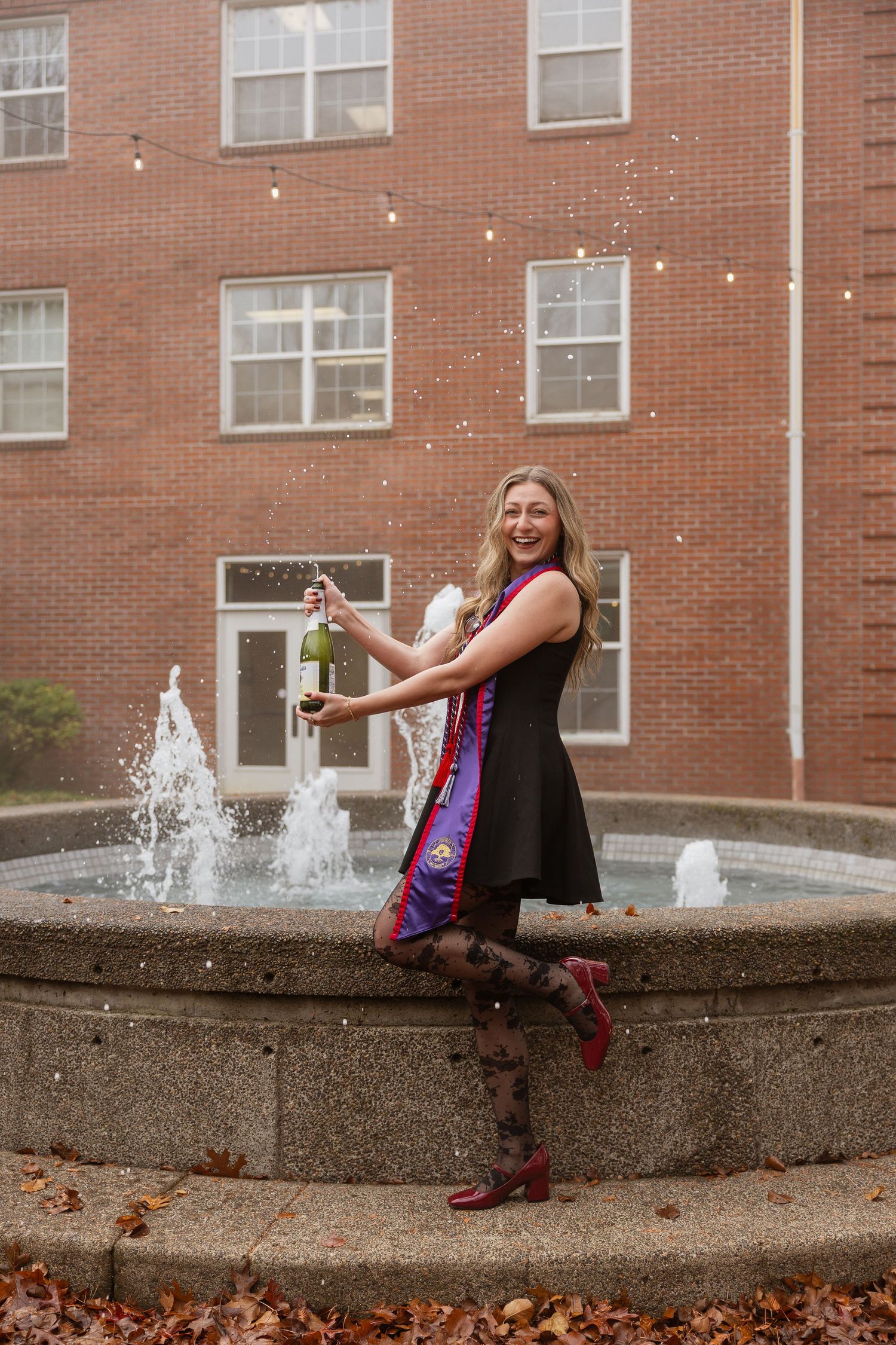 A person in a black dress and red boots poses playfully by a fountain on a brick building's courtyard during autumn.