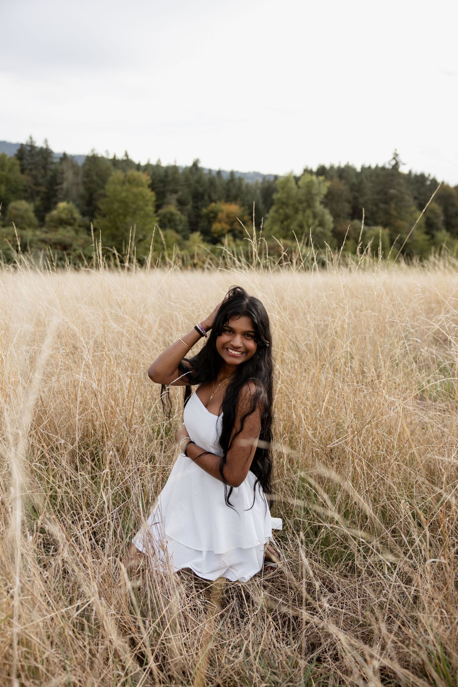 A person in a white dress sits in a golden field of tall grass during sunset with trees in the background.