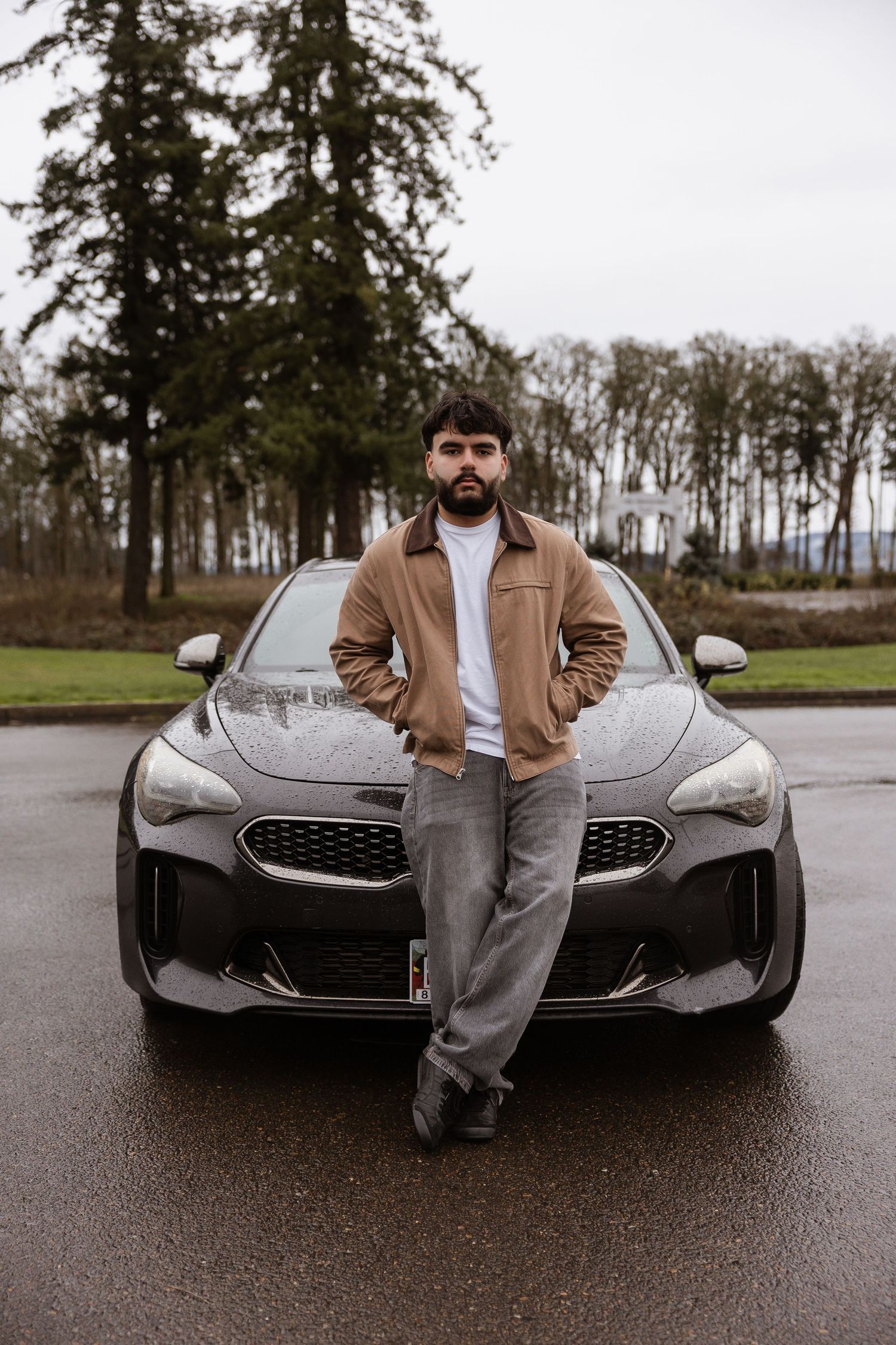 A person in a tan jacket poses with a sleek silver luxury car on a wet asphalt road near trees.
