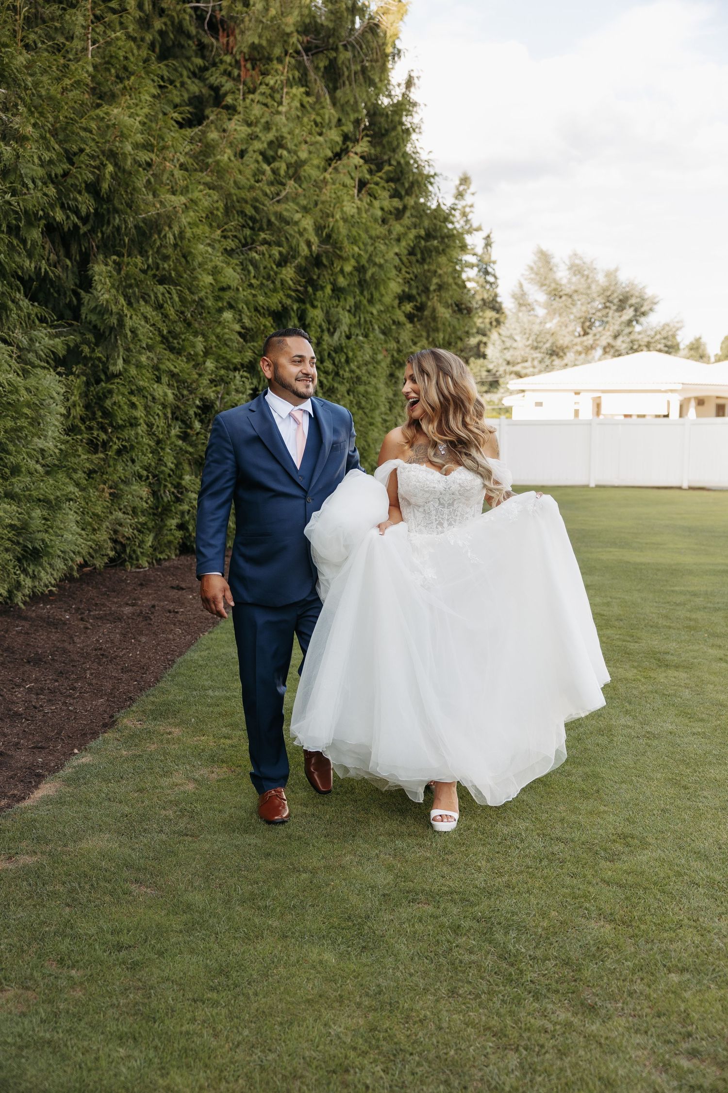 A couple walks together on a lush green lawn with white wedding attire and navy suit against a hedge backdrop.