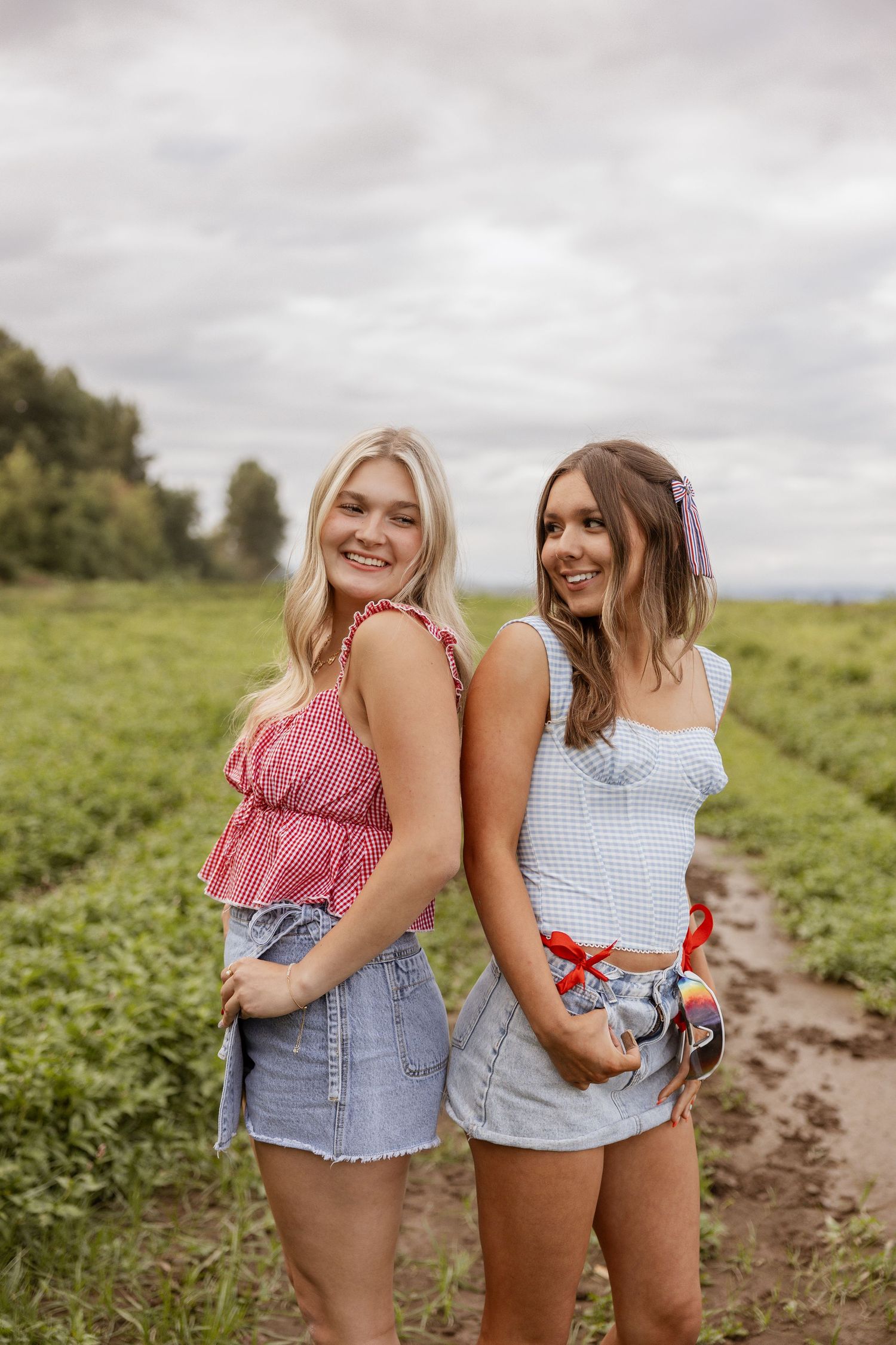 Two friends pose back-to-back in summer outfits while standing in a green field on a cloudy day.