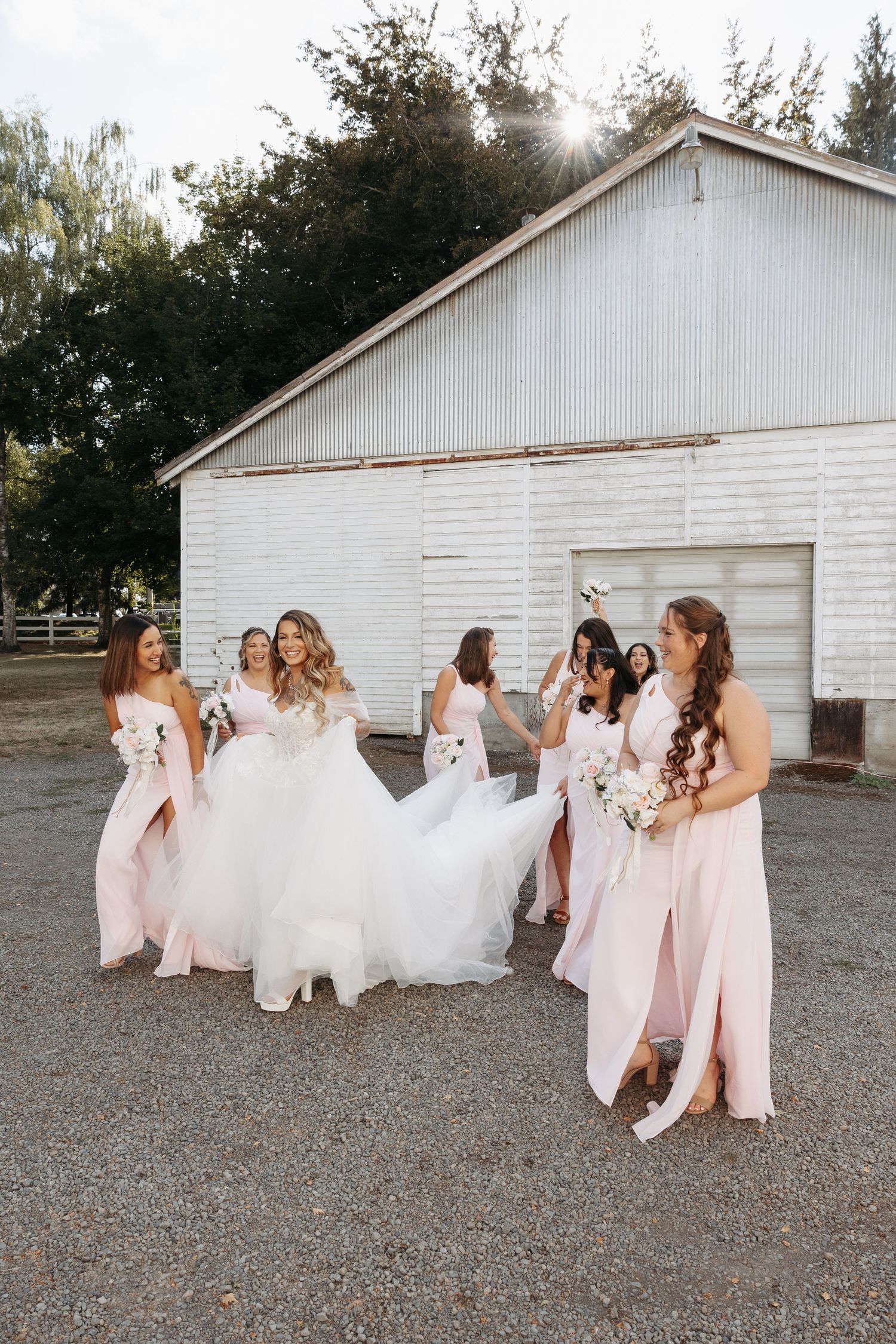 Bridesmaids in pink dresses laughing together outside a white rustic barn.