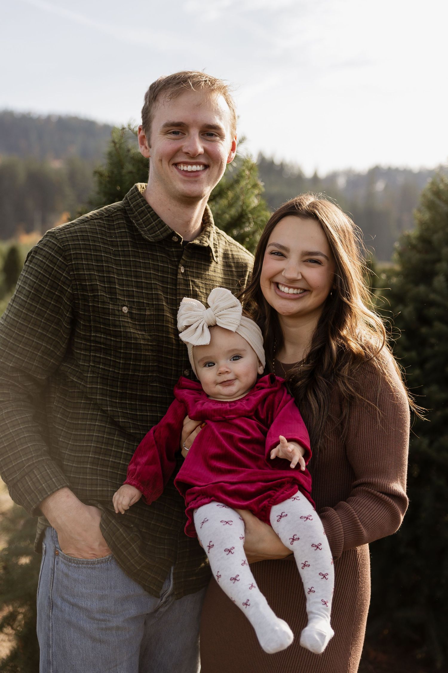 Young couple smiling outdoors at a Christmas tree farm, holding their baby girl in a red dress and bow headband.