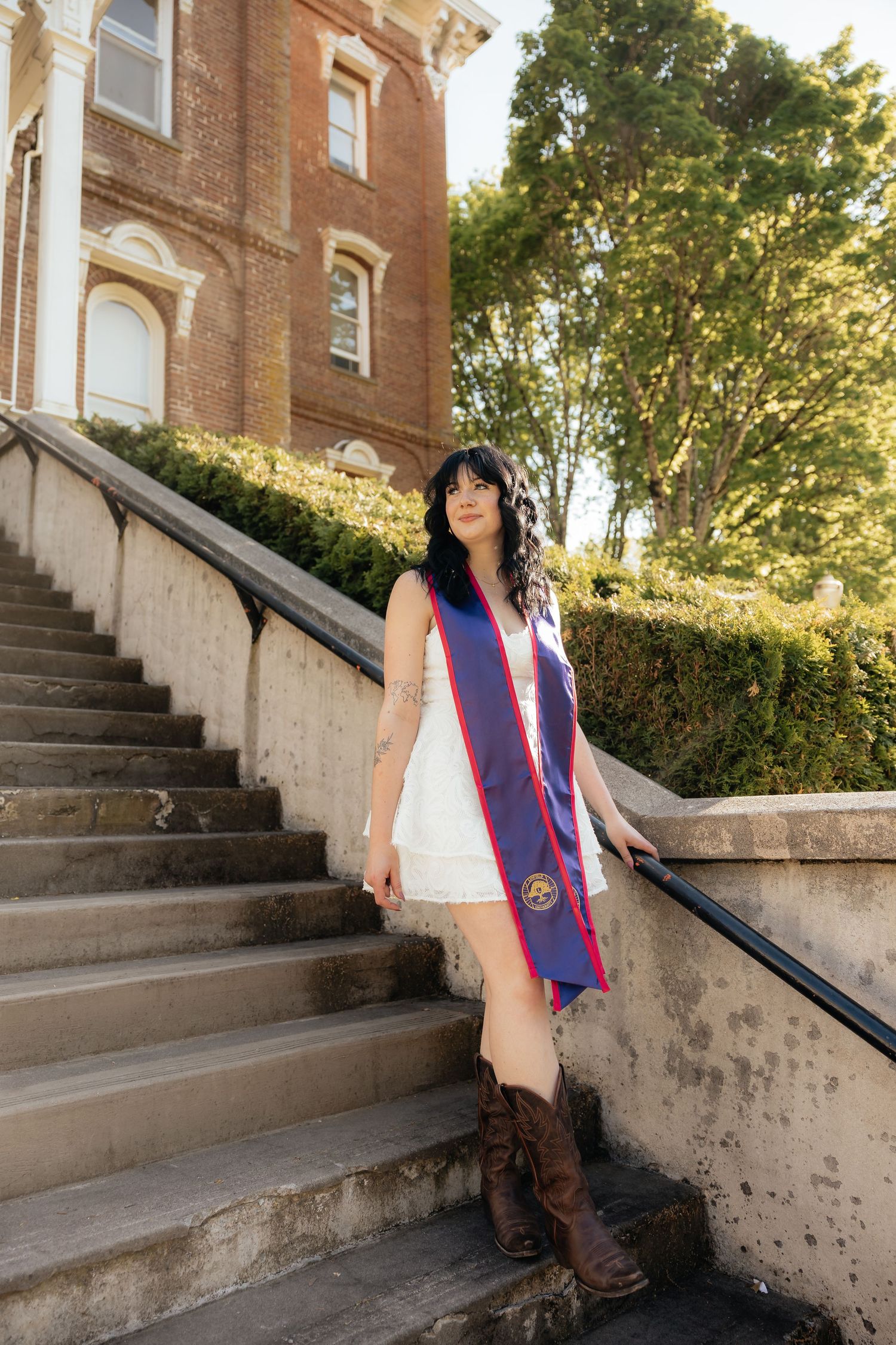 Graduate in white dress and navy sash poses on stone steps outside brick university building.