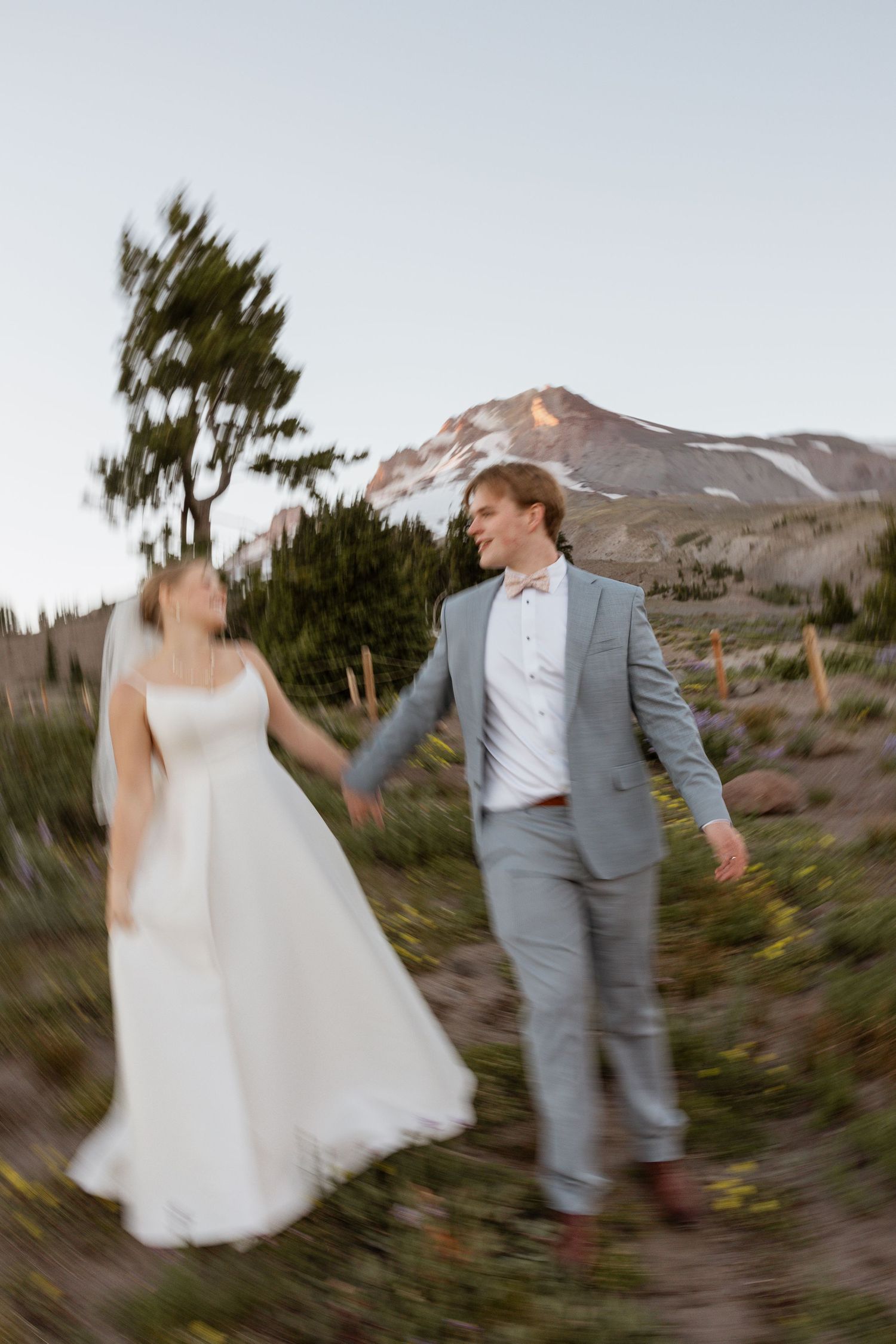 A couple in wedding attire dances and twirls together against a mountainous desert landscape at sunset.