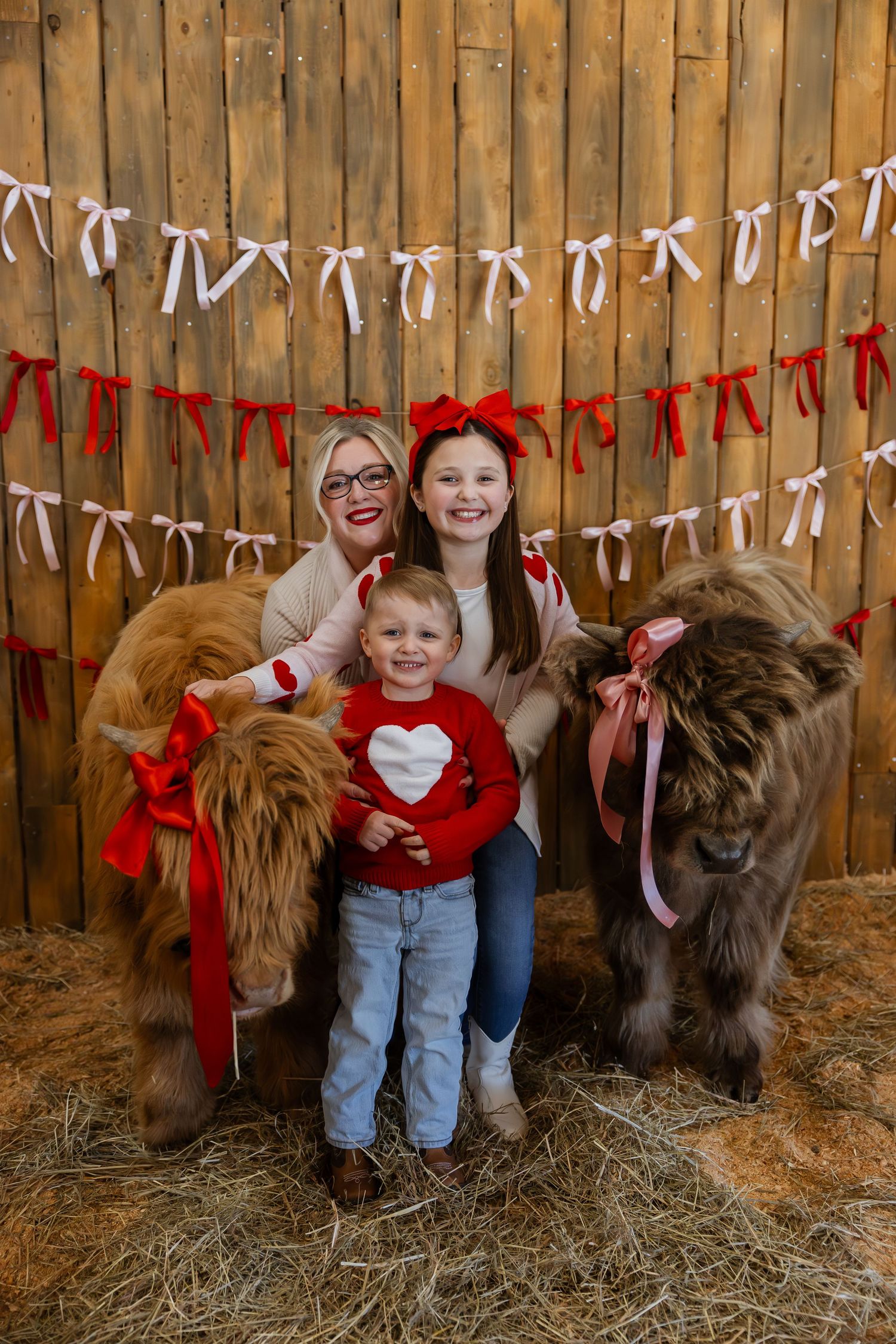 A family poses with miniature ponies in a barn decorated with red and white ribbons for holiday photos.