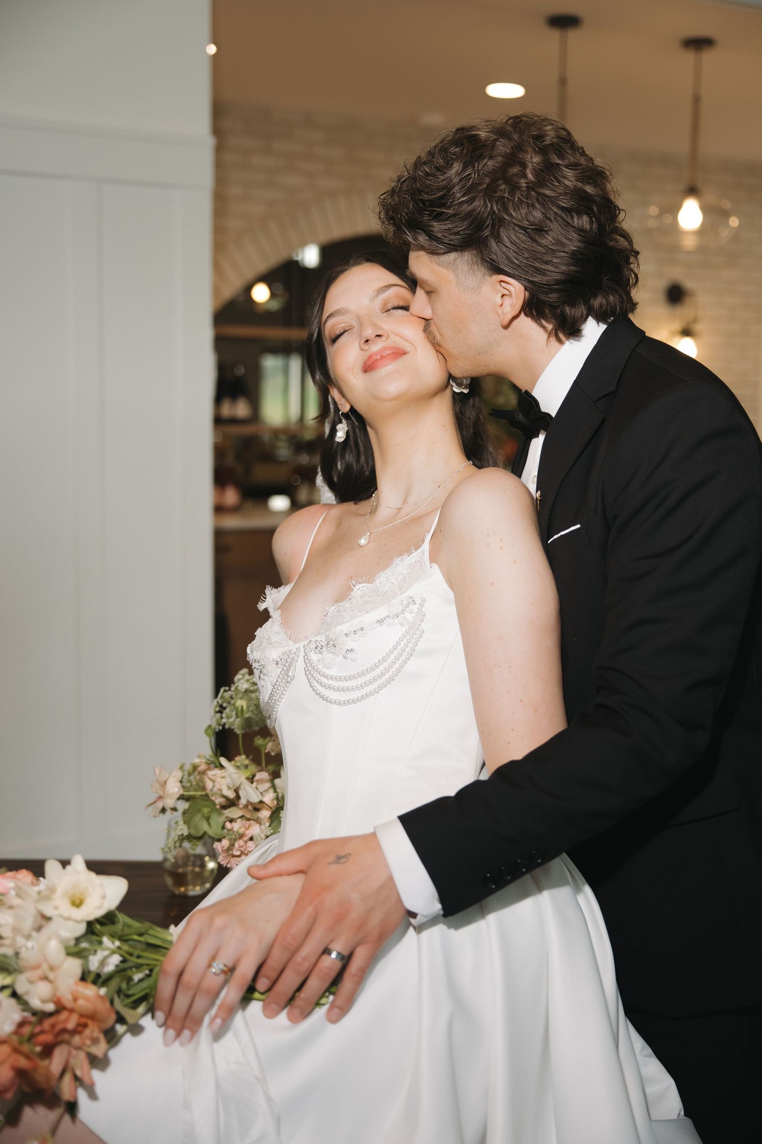 Groom in black tuxedo kissing bride in white wedding dress, surrounded by floral arrangements indoors.