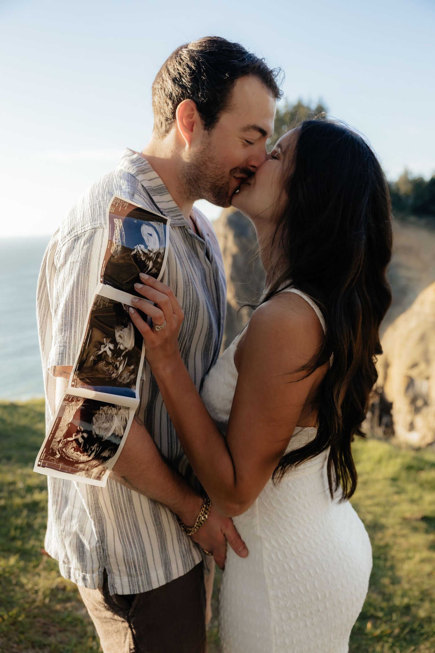 Couple kissing outdoors holding ultrasound photos, woman in white dress showing baby bump, ocean cliffs in background.